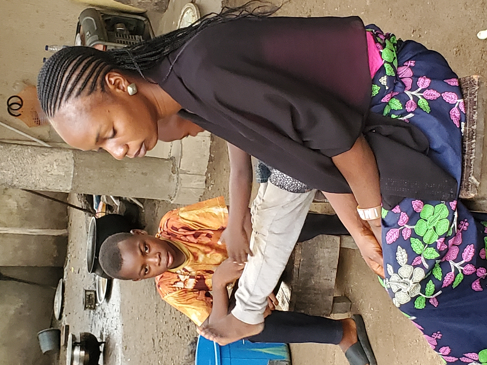 In Mararaba, Nigeria, a woman with braided hair and wearing a black shirt and a blue floral skirt is attending to a young boy. The boy, dressed in a yellow and orange patterned shirt and black pants, is seated on a blue bucket. The woman is holding his leg, possibly applying something or checking it. The setting appears to be an outdoor or semi-outdoor space, likely a backyard or courtyard. Various cooking utensils and pots are scattered nearby, indicating a domestic environment. The overall impression is of a scene where someone is providing care or assistance to a child in a simple, perhaps rural or semi-urban setting.