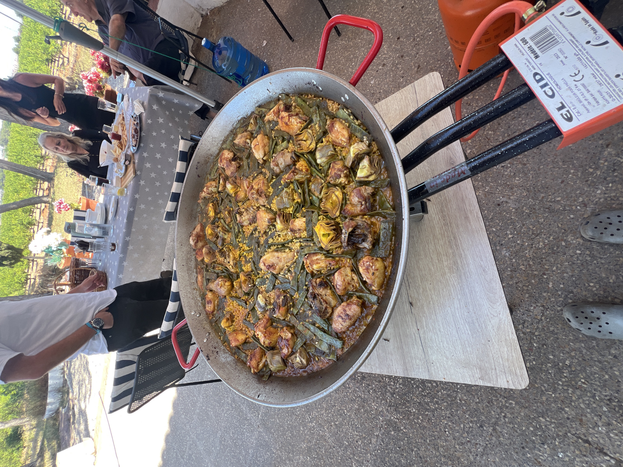 Here is a forensic description of the image:
**Scene**: An outdoor paella preparation event.
**Location**: Museros, Spain. This is determined by the text on the EL CID propane tank label, which indicates "44900 TORRENT VALENCIA SPAIN". Torrent is near Valencia, which is near Museros.
**Subjects**:
* A large paella pan filled with paella. The paella contains rice, meat (possibly chicken), green beans, and artichoke hearts.
* A paella burner stand connected to a propane tank.
* People in the background, including those sitting at a table with food and drinks.
* One person wearing gray Crocs is partially visible.
* One person in a white shirt is partially visible.
**Objects**:
* Table covered with a star-patterned tablecloth.
* Various drinking glasses and bottles on the table.
* A wooden board is placed beneath the paella burner stand to protect the ground.
* Propane tank with an "EL CID" label.
**Actions/Interactions**:
* The paella is being cooked outdoors.
* People appear to be gathered for a meal or event.
**Environment**:
* The event is taking place outdoors on a paved surface.
* There are chairs and tables set up for dining.
* Green foliage visible in the background, potentially vineyards.