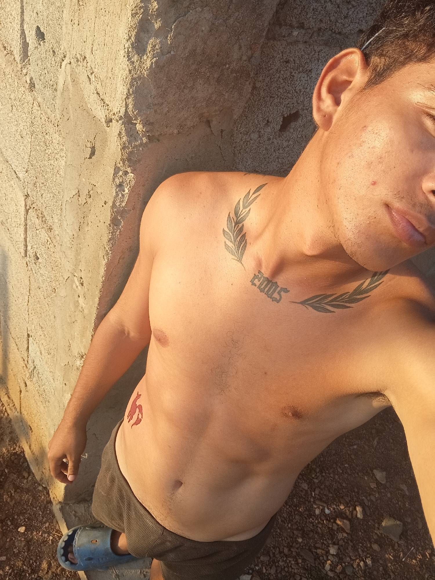 A shirtless young man stands outside next to a concrete wall. The location may be Altagracia, Venezuela. The man has a tattoo on his chest that says "2005" and a laurel wreath tattoo around it. He also has a tattoo of a red bird or flame on his lower abdomen. He is wearing dark shorts and blue sandals. The ground is gravel.
