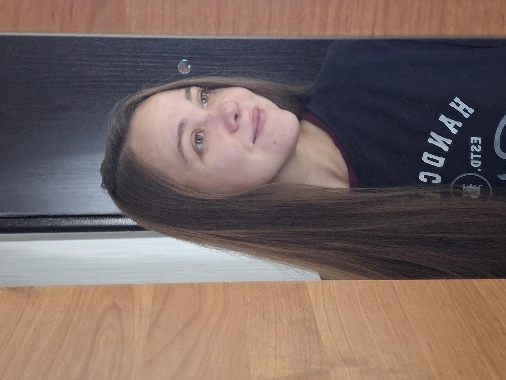 The image shows a young woman with long, brown hair and light eyes, looking to the right. She is wearing a dark blue t-shirt with white lettering that appears to be in Russian. The background consists of a doorway with a dark wooden frame on the right and a light-colored door on the left, with a narrow vertical strip of light gray between the wood and the door. There is a circular, reflective element on the dark door. The woman is positioned to the left of the center of the frame, with her face and upper torso visible. A portion of a wooden surface is on the far left edge of the image. The scene appears to be indoors, possibly in a residential setting, and there are no elements that specifically place it in Chaikovsky, Russia, other than the prompt's instruction.