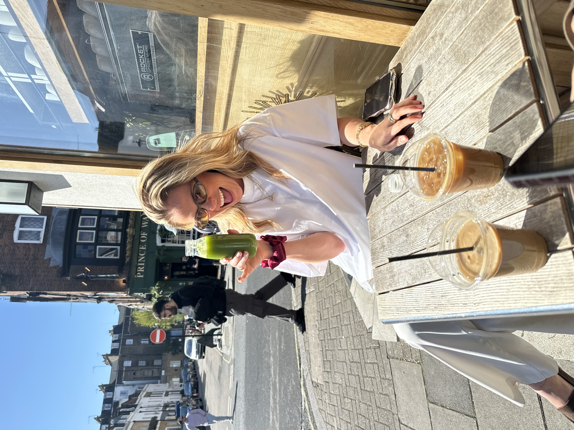 The image captures a woman sitting at a wooden table outside a café, holding a green juice bottle in her left hand and smiling at the camera. She is wearing a white shirt and glasses. Two iced coffees with straws are on the table. A mobile phone is also visible on the table.
Through the window behind her, the reflection of a building across the street reads "PRINCE OF WALES". In the background street scene, there's a street with cars and pedestrians. A cyclist is passing by. A no-entry sign is visible on the street. The buildings suggest a residential area within London. The clear sky and sunlight indicates it is daytime.