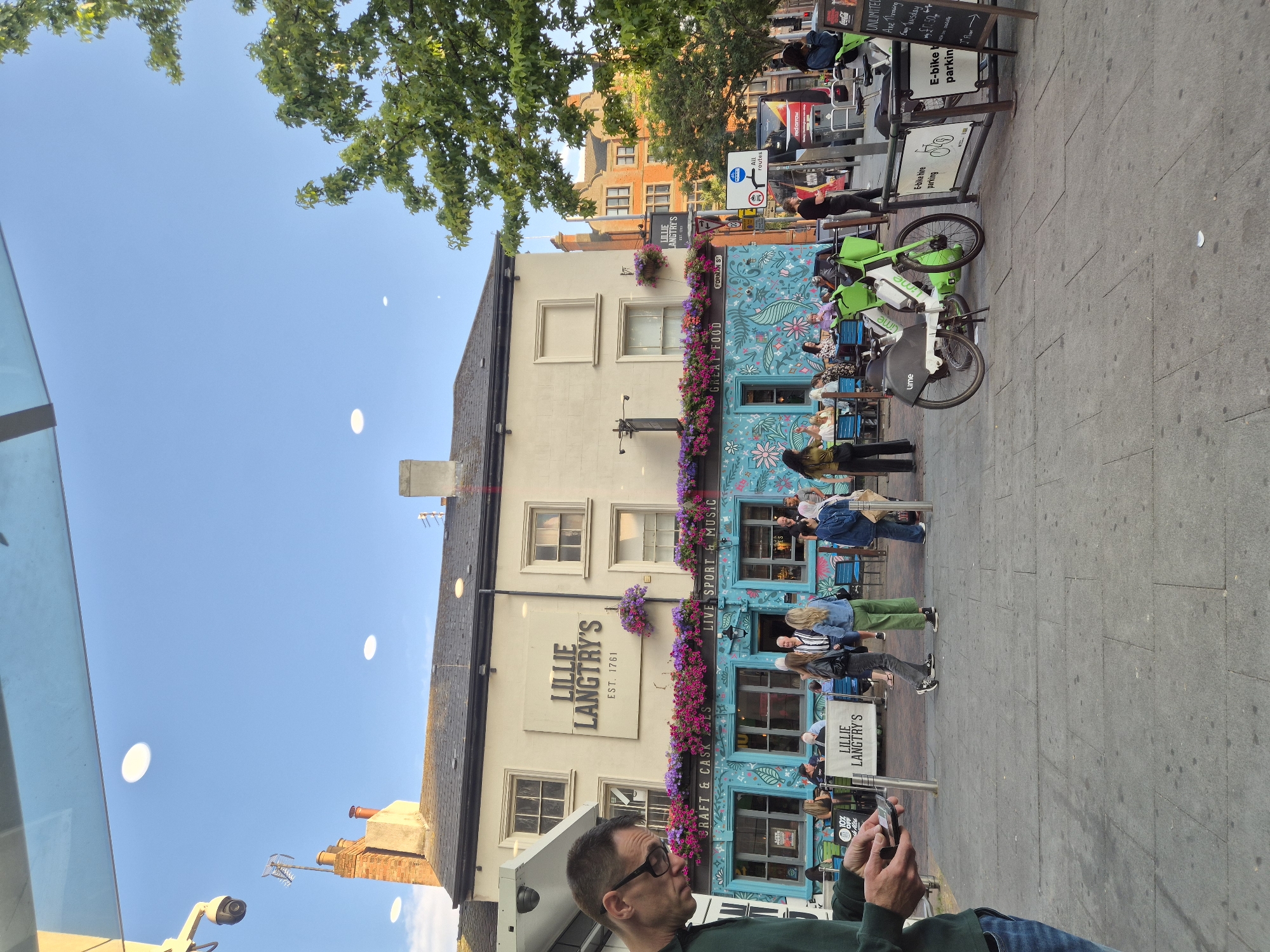 The image shows the exterior of Lillie Langtry's pub, located on Forman Street in Nottingham, United Kingdom. The pub has a distinctive turquoise facade adorned with floral murals and is decorated with hanging baskets of flowers. The pub's name and "EST. 1761" are prominently displayed on its cream-colored upper story. Several people are visible sitting at tables outside the pub, and others are walking on the pavement. In the foreground, a man wearing glasses and a green top is looking down at his phone. Two green electric bikes, branded with "Lime," are parked on the pavement near a sign indicating "E-bike hire parking." The scene is set against a blue sky with scattered clouds, and a tree with green foliage is visible to the right. The overall atmosphere appears to be casual and daytime.