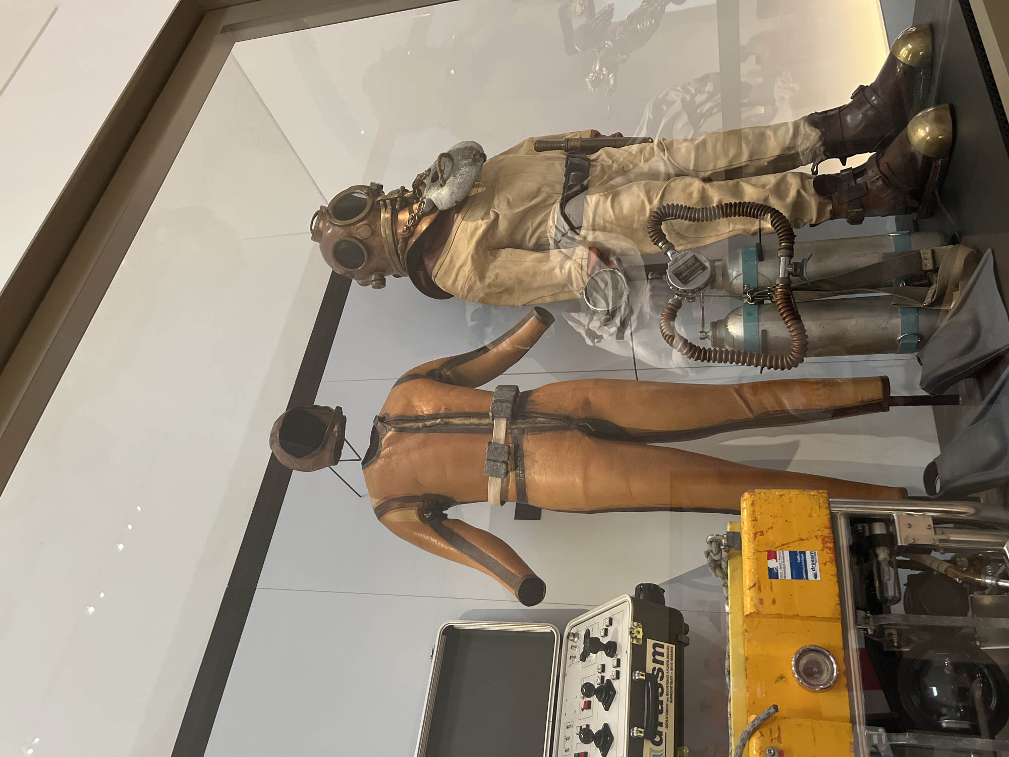 The image displays a museum exhibit featuring vintage diving equipment, likely located in Paris, France, possibly at the Cité des Sciences et de l'Industrie. On the left is a tan diving suit with a hood, displayed on a mannequin. To its right, a more elaborate copper helmet diving suit is also presented on a mannequin, complete with a mask, hoses, and what appear to be two air tanks. In the foreground, to the left of the diving suits, is a yellow equipment box with various controls and a black briefcase-style case. The exhibit is viewed through glass, reflecting some of the surrounding environment.