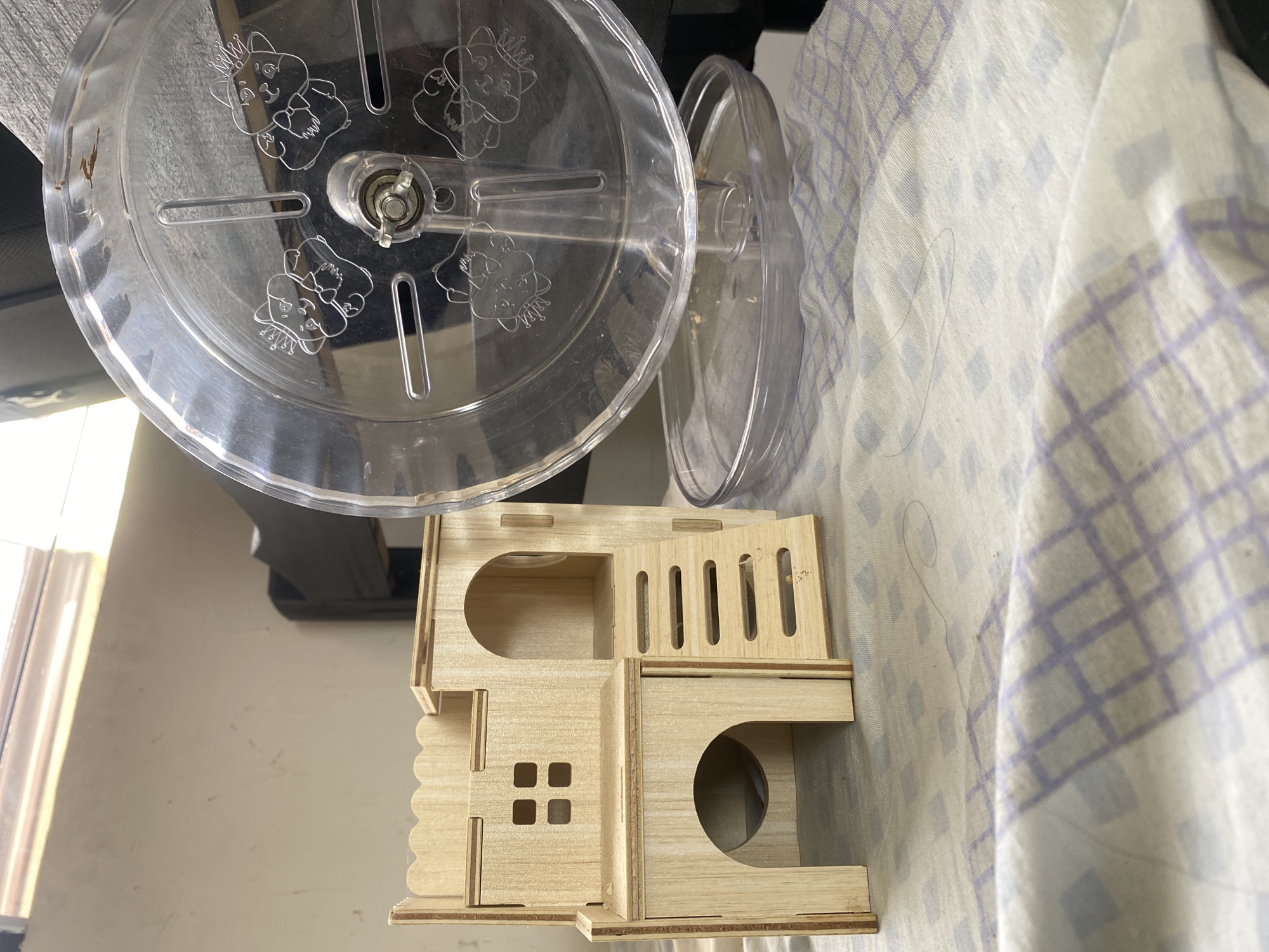 The image shows a wooden hamster house and a clear plastic hamster wheel placed on a white fabric with a purple grid pattern. The wooden house has multiple levels, stairs, and openings, resembling a miniature castle. The hamster wheel has cartoonish animal faces engraved on it and a small tray at its base. The background is blurred but suggests an indoor environment with a window and some dark furniture. The scene is set in a room, likely a home, on a fabric surface. There is no indication of the specific location in Davao City, Philippines.