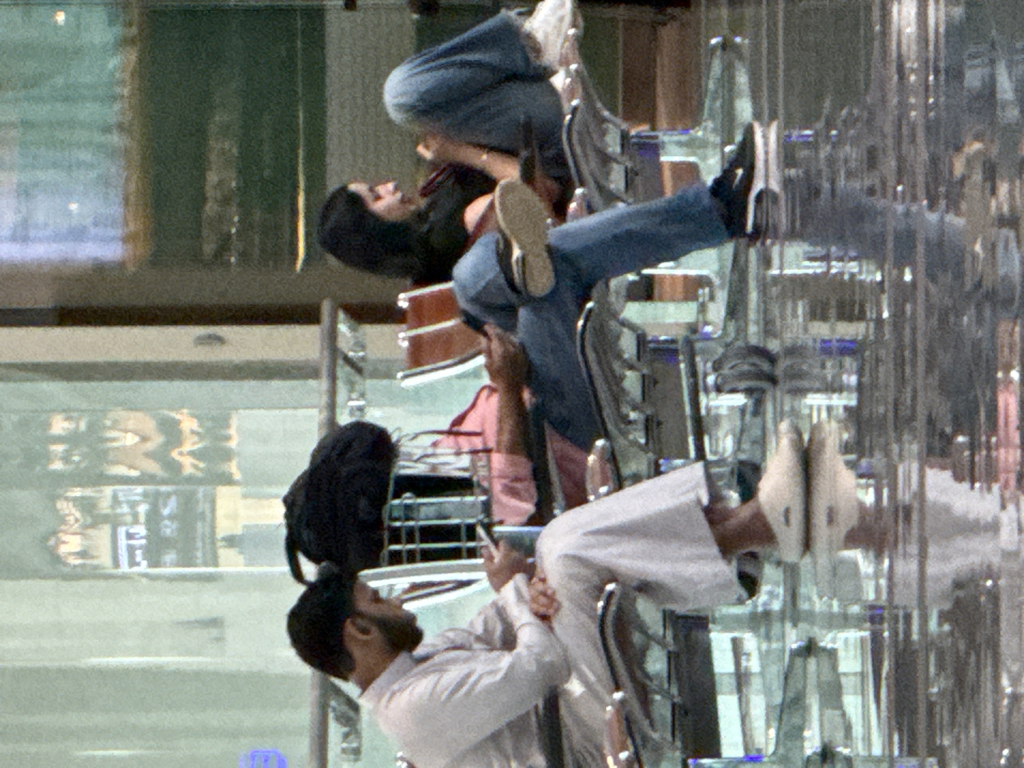 The image depicts a scene likely within an airport terminal in Seeb Province, Oman. Two individuals are seated in adjacent rows of airport seating, facing away from the viewer. On the left, a man wearing a white thawb and headdress is looking down at his mobile phone, which he holds in his right hand. His left hand rests on his lap. Beside him, a black backpack is placed on his seat. To his right, a woman with dark hair, wearing a black top and blue jeans, is seated with her legs crossed and her head tilted down as she also appears to be looking at a mobile device. The floor is highly reflective, suggesting a polished surface, possibly marble or tile, with reflections of the ceiling lights and the seating area visible. In the background, glass partitions and seating structures are visible, indicative of a public transit waiting area. The overall lighting is bright, with artificial illumination.