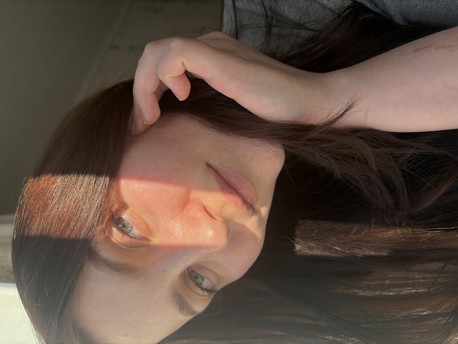 A woman's face is partially shadowed by sunlight. She has light skin, green eyes, and brown hair, with her hand near her forehead. The image is rotated. Part of her arm is visible, with faint marks on the skin. She is wearing a gray top. The light creates a sharp contrast on her face.