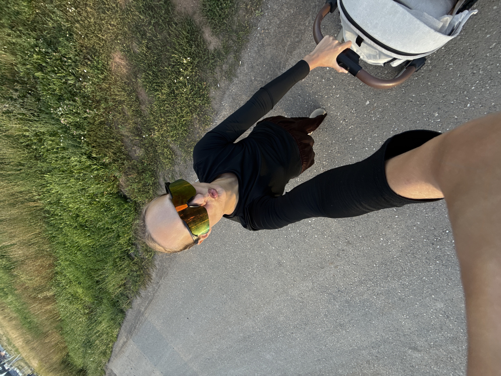 A person is taking a selfie while pushing a stroller. They are wearing black long-sleeved shirt, brown pants, and reflective sunglasses. The person is on an asphalt path next to a grassy area with some small white flowers. The stroller is light-colored with a brown handle. The image appears to be taken outdoors during the day.