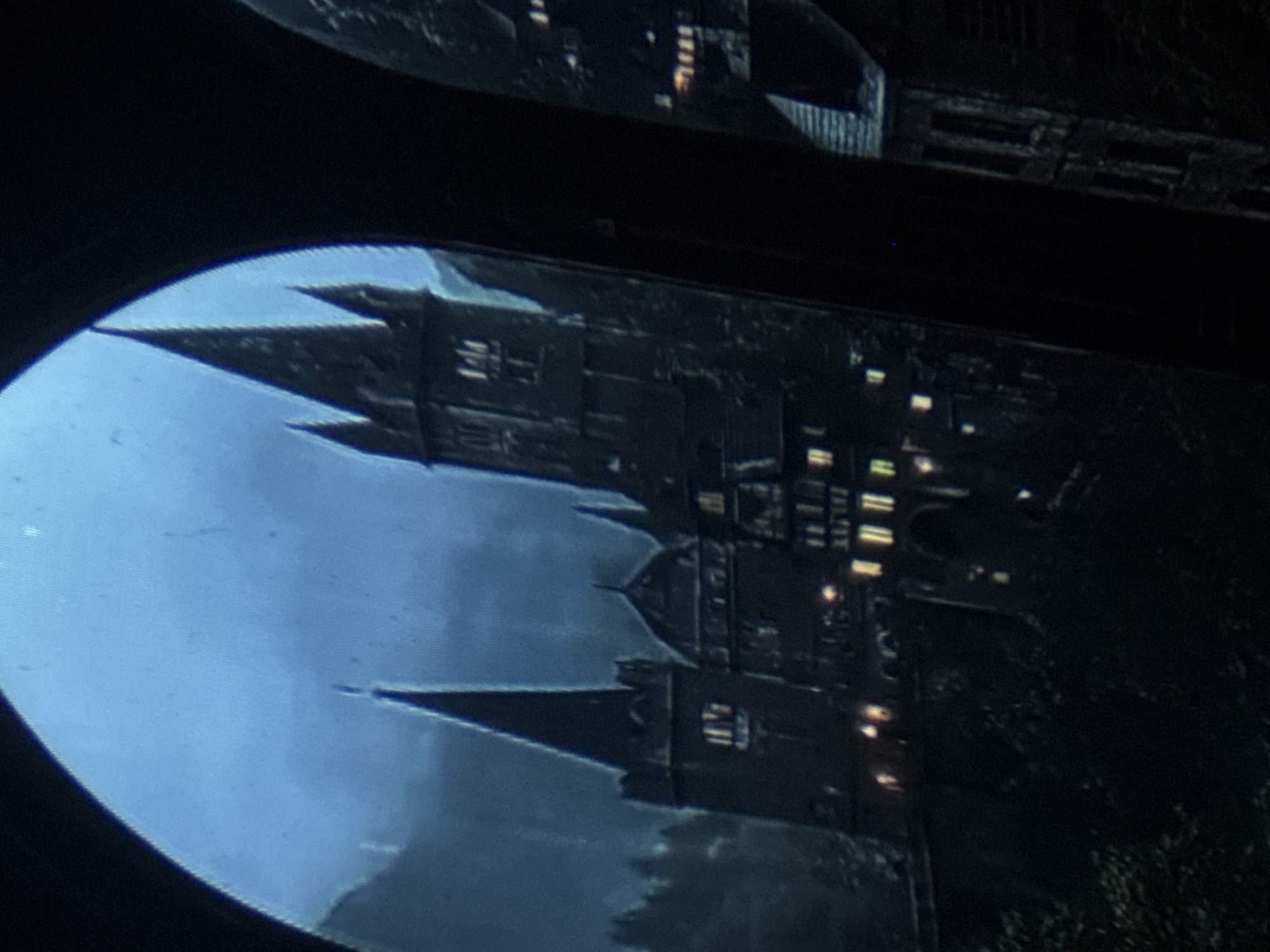 The image depicts a nighttime view of a castle-like structure, possibly Hogwarts, seen through an arched opening. The castle is illuminated by numerous lit windows, and its imposing spires and towers are silhouetted against a dark, cloudy sky. The surrounding environment appears to be mountainous or forested, with hints of trees and rugged terrain visible in the foreground and background. The overall impression is one of a dramatic and somewhat mysterious scene. There is no indication of the city of Tbilisi, Georgia, in this image.