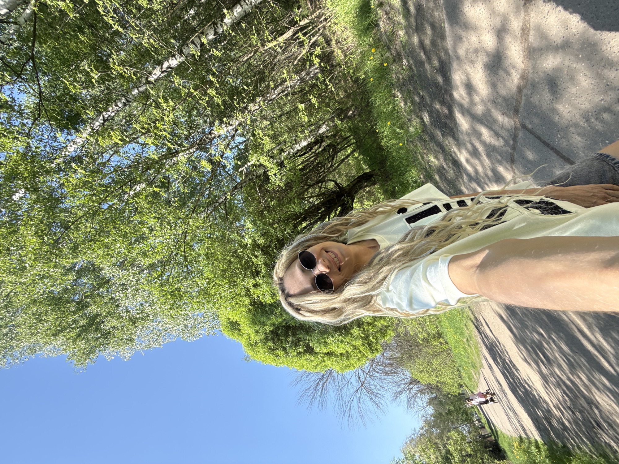 A selfie taken outdoors in Rusanovo, Russia shows a blonde woman with sunglasses lying on the ground. Above the woman are trees with green leaves against a blue sky. To the right of the woman is a paved path with dappled sunlight. In the distance, a stroller is visible on the path. The woman is wearing a light-colored shirt. Part of her leg with gray shorts is visible in the top right corner of the image.