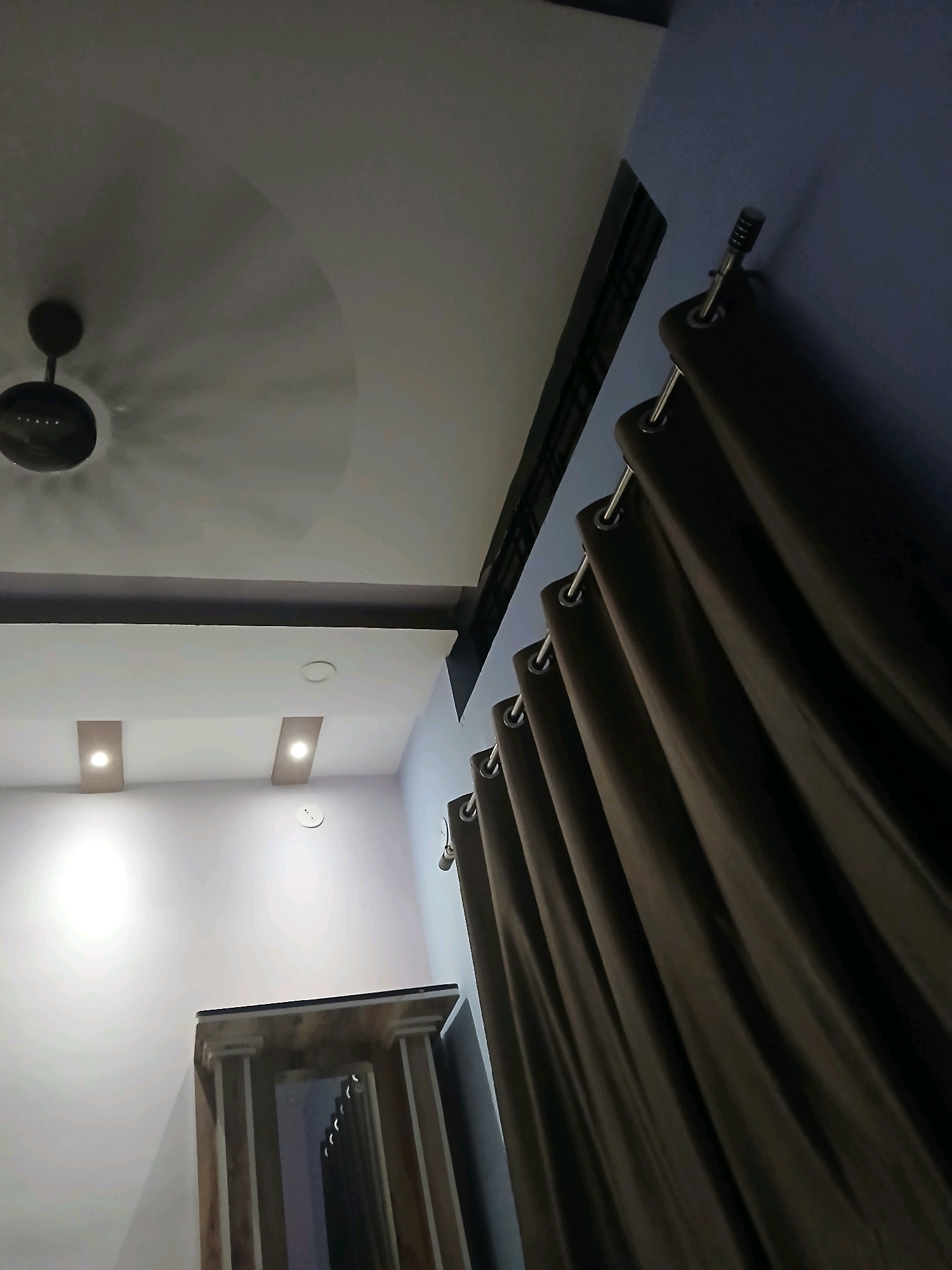 The image depicts an indoor scene, likely a room. In the upper left, a dark ceiling fan is visible against a white ceiling. Moving towards the right, a section of a dark ventilation grill is present. Below this, a dark brown curtain hangs from a rod with metal grommets. To the left of the curtain, there is a white wall with two rectangular recessed light fixtures emitting a warm glow. Further left, a circular recessed light is also visible. In the lower portion of the image, a decorative wooden structure, possibly a doorway or furniture, is partially visible. The overall lighting suggests it is evening or the lights are dimmed. No people or specific city landmarks of Ambikapur, India are identifiable in the image.