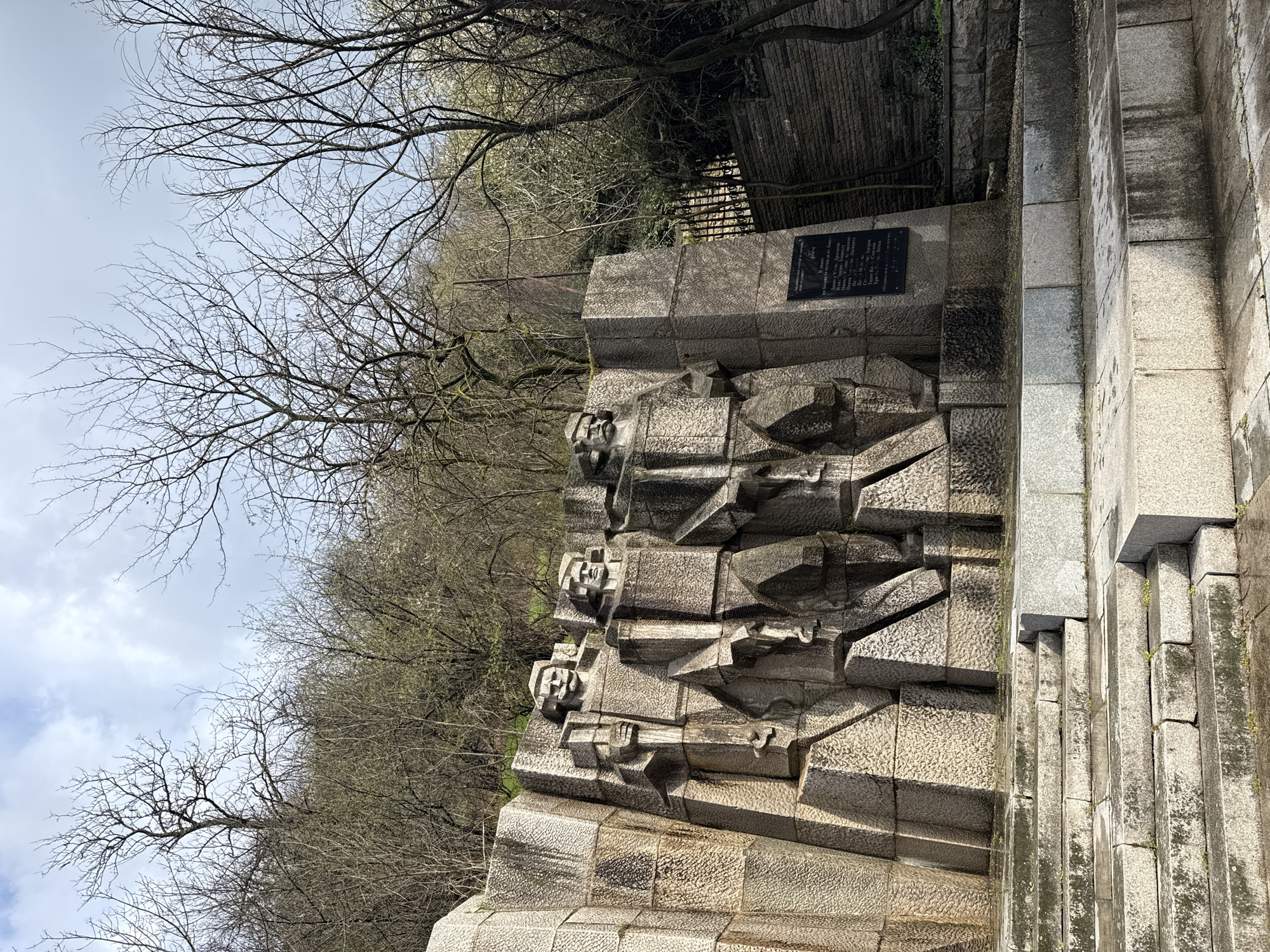 The image features a stone monument in Lovech, Bulgaria. The monument's facade is constructed from rectangular blocks, featuring sculpted faces and the suggestion of rifles, possibly representing soldiers. A black plaque with inscription is affixed to the upper portion of the monument. Bare trees and a cloudy sky form the backdrop. The monument is located on the edge of a walkway or road surface that is paved with stone.