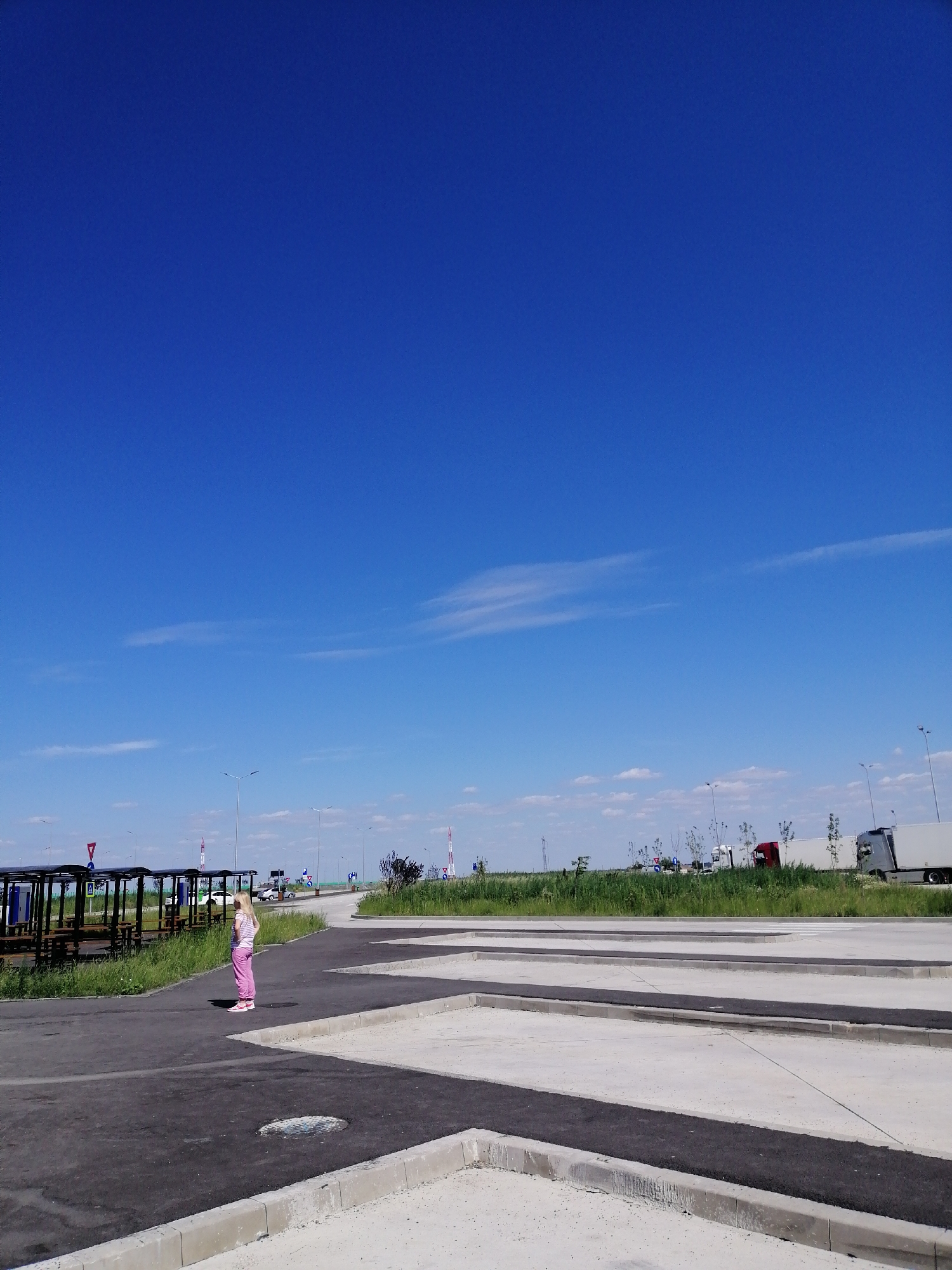 A young girl with blonde hair is standing on a paved surface, wearing a pink top and pink pants. She is facing away from the camera, looking towards a row of covered picnic tables with wooden benches. In the background, there are several large trucks parked along a grassy area. The sky is a vibrant blue with a few wispy clouds. The scene appears to be a rest stop or a parking area, possibly near a highway.
