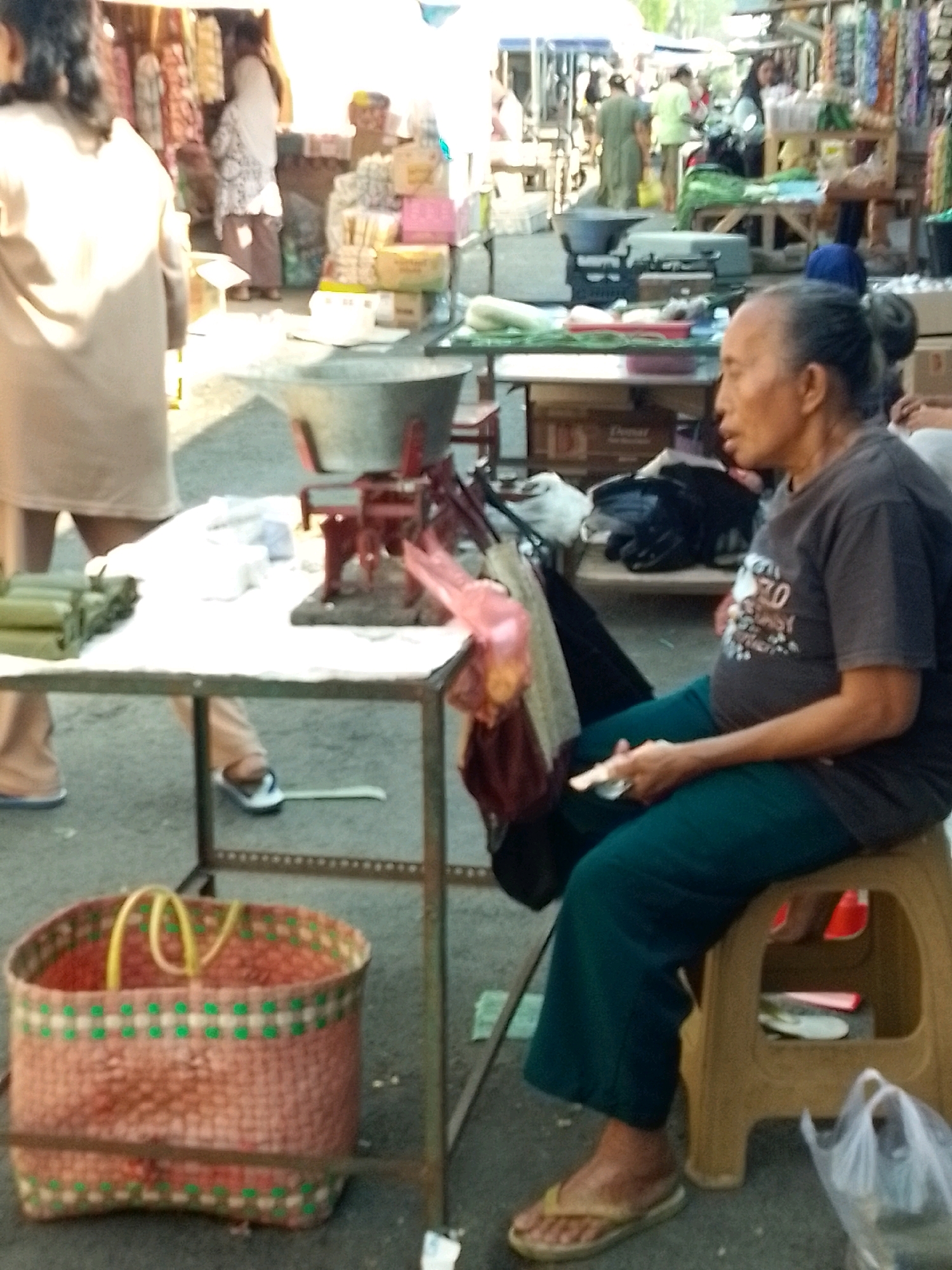 A middle-aged Indonesian woman is sitting on a yellow stool in an outdoor market, likely in Jember, Indonesia. She is wearing a dark t-shirt and teal pants, with flip-flops on her feet. She is holding a small, flat object in her right hand. The market appears busy, with other people visible in the background, as well as stalls selling various goods. In the foreground, there is a large woven basket with yellow handles. To the left of the woman, a table is set up with some food items and a scale. The overall scene depicts a typical market environment in Indonesia.