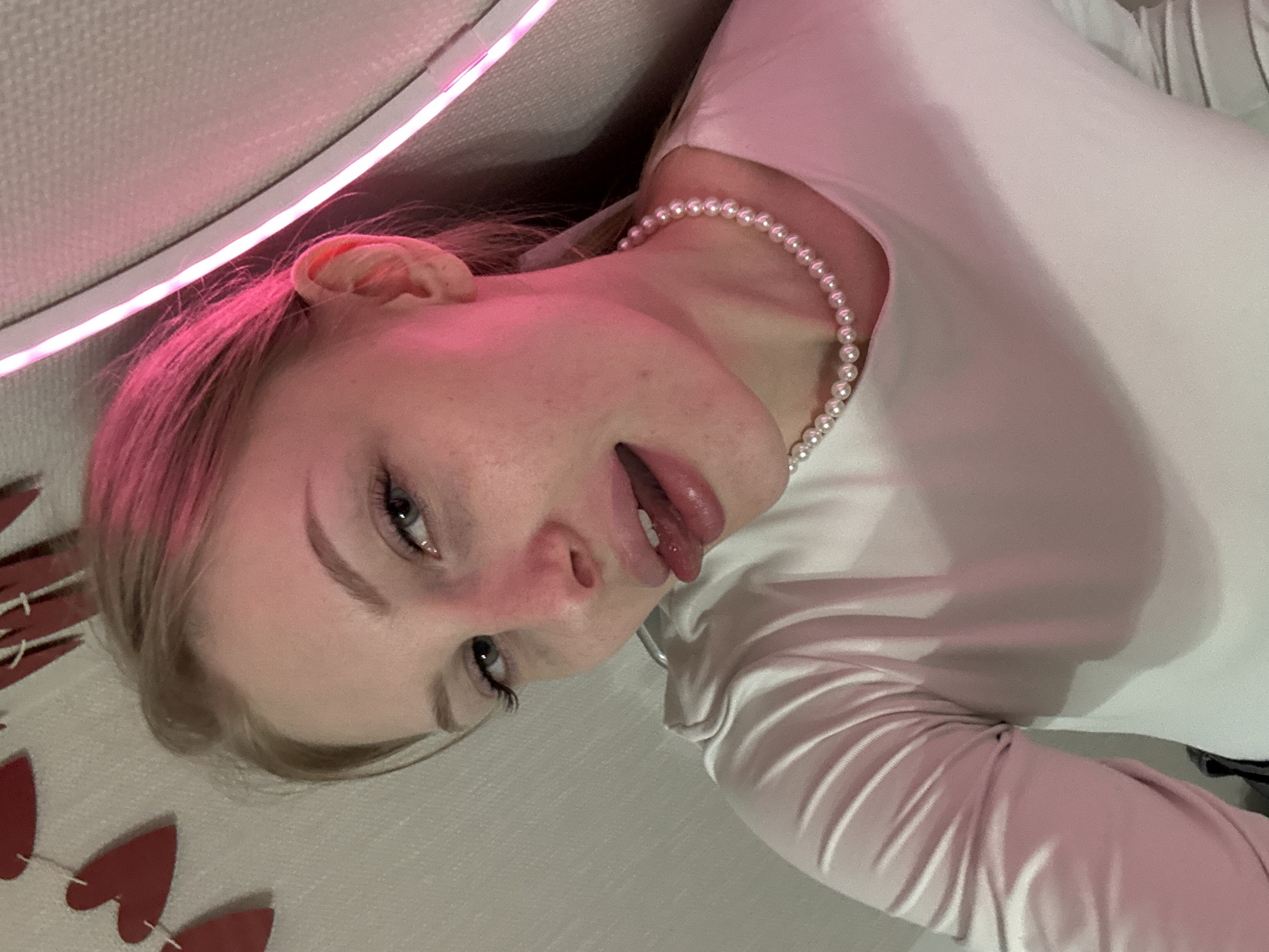 A young woman is depicted in a close-up, chest-up shot, positioned diagonally within the frame. She has light skin and light hair that appears dyed or strongly lit with a pink hue. She is wearing a white long-sleeved top and a multi-strand pearl necklace. Her head is tilted, and she looks directly towards the viewer with her eyes open. Her mouth is slightly open, with her tongue visible, suggesting a playful expression. Behind her, a strip of glowing pink or magenta LED light is visible, casting a strong, warm glow on her hair, ear, and face. The background appears to be an indistinct indoor setting, possibly a wall or upholstered furniture. In the lower left corner, several red heart-shaped decorations, possibly part of a garland, are visible. There is no visible text in the image.