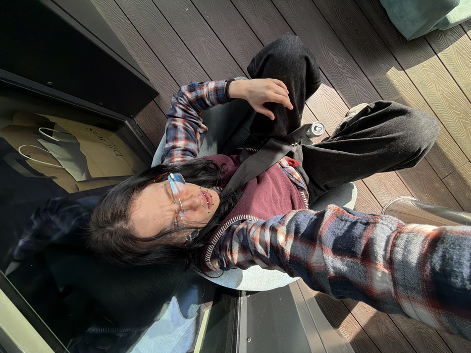 An individual with long, dark hair, wearing rimless glasses with blue-tinted lenses, sits in an outdoor or semi-outdoor commercial setting in Sofia, Bulgaria. The person, who has multiple facial piercings (including on the nose bridge, perioral region, and ear gauges), is clad in a blue and red plaid flannel shirt over a maroon vest and dark pants. They are seated in a light-colored chair with visible casters, looking directly at the camera, which is held in their outstretched right hand. The individual's eyebrows are furrowed. A black strap crosses their chest. To the bottom left, a clear beverage container is partially visible near the right arm, and a silver beverage can rests on the floor next to the chair. The flooring consists of light brown composite decking planks. A dark, reflective vertical surface on the left side of the image displays reflections of two light-colored paper shopping bags, one clearly branded "DILIOS," indicating the location is within a commercial area, likely a shopping mall, in Sofia. Strong natural sunlight illuminates the scene, creating distinct shadows on the decking.