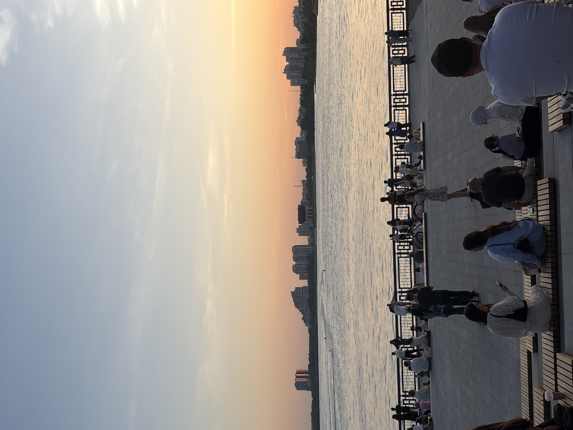 The image captures a sunset scene in Kazan, Russia. A river divides the frame, with the city skyline silhouetted against the orange and yellow hues of the setting sun on the left. On the right bank of the river, a promenade is lined with a black metal railing and benches. Many people are sitting and standing along the promenade, watching the sunset and interacting with each other. Some individuals are using mobile phones, while others are seated on the benches, taking in the view. The lighting suggests it is either early morning or late evening.