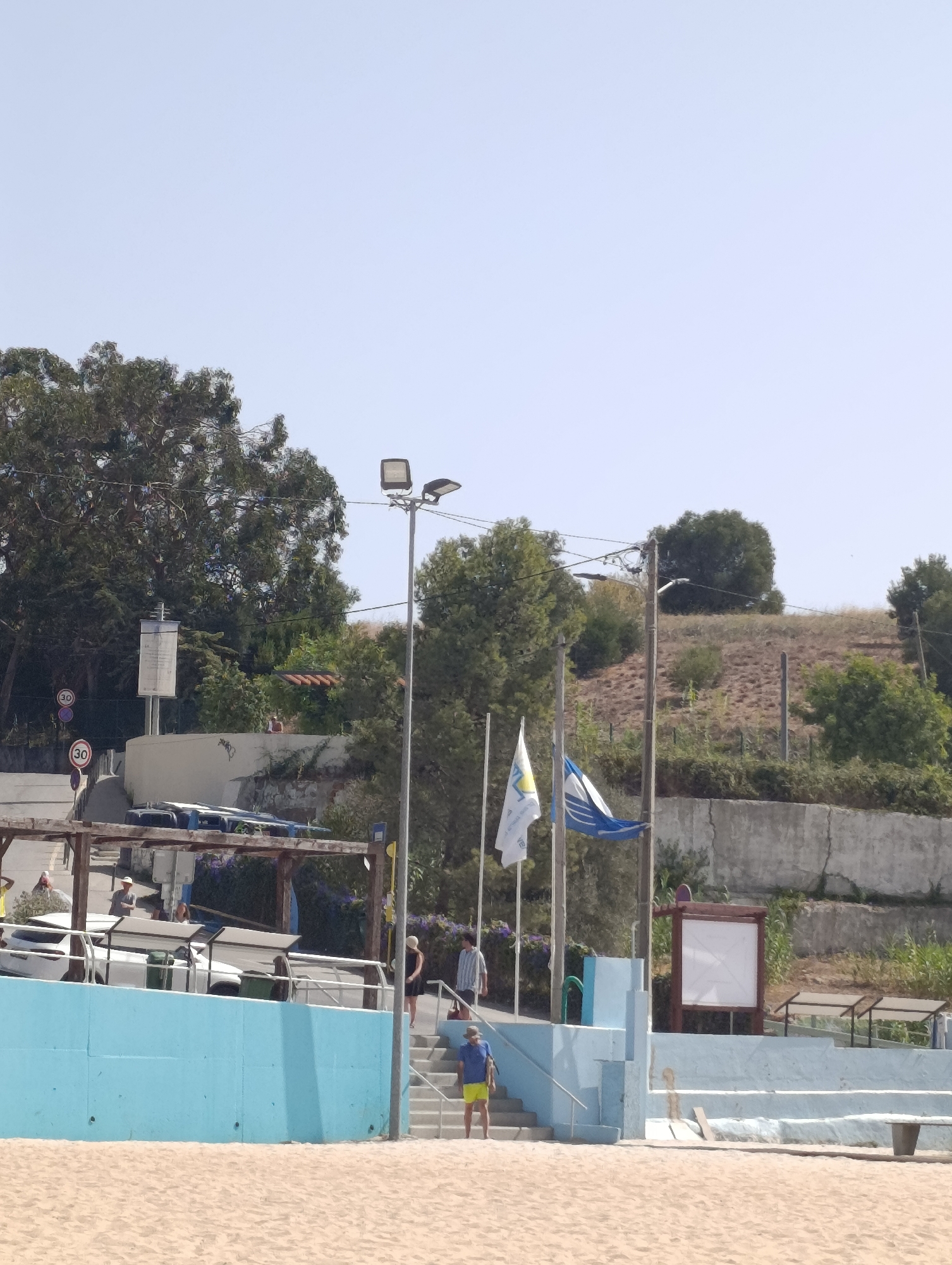 The image captures a sunny day at a beach in Setúbal, Portugal, likely near the Avenida Luísa Todi waterfront. In the foreground, a sandy beach stretches across the bottom of the frame. A bright turquoise wall defines the edge of a raised area, behind which are steps leading up. On these steps, a person wearing a blue shirt and yellow shorts stands facing left. Further up the stairs, to the right, another person in a dark dress and a person in a striped shirt are visible.
Above the turquoise wall, a wooden pergola covers a row of parked vehicles, including white cars and what appear to be trash bins. A prominent light pole with two lamps stands to the right of the frame, with flags flying nearby. One flag is white with blue and yellow emblems, and another is blue and white. The background features a steep, vegetated slope with a concrete retaining wall. In the distance, trees and sparse vegetation cover the hillside under a clear blue sky. Traffic signs indicating a speed limit of 30 are visible on a pole to the left.