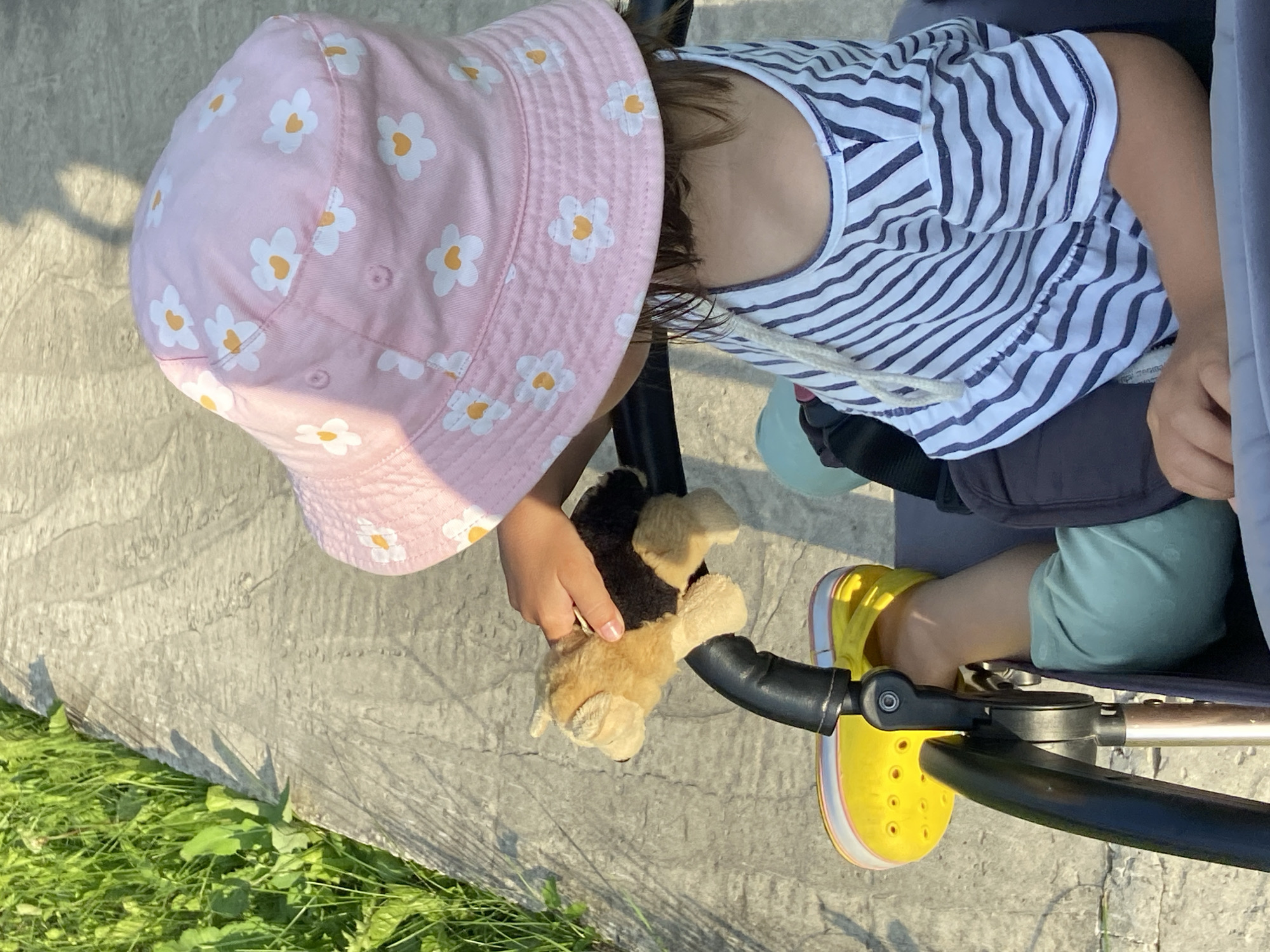 An overhead, close-up shot captures a young child wearing a pink bucket hat with a daisy pattern and a blue and white striped t-shirt. The child is seated in a stroller, holding a small, tan and black plush toy dog. Their left foot, clad in a yellow Croc-style shoe, rests on the stroller's frame. The stroller and child are positioned on a concrete path adjacent to a grassy area. The lighting suggests it is daytime, with shadows visible on the path. The scene is generic and does not provide any specific visual cues to indicate its location in Tyumen, Russia.
