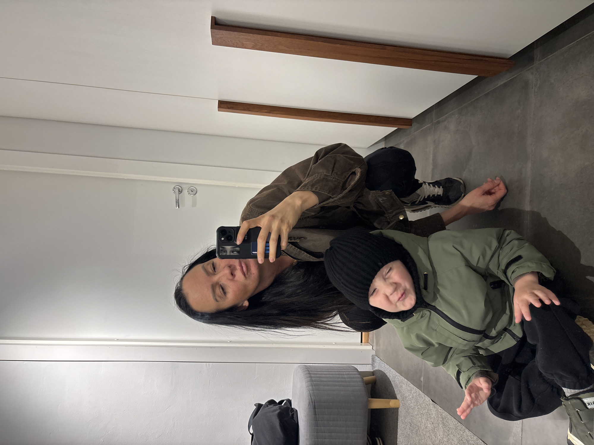 A selfie shows a woman and a young child, presumably reflected in a mirror. The woman has long, dark hair and is wearing a brown jacket. She is holding a smartphone and taking a picture of herself and the child. The child is wearing a black beanie and a green jacket, and is also visible in the frame. Behind them is a bathroom setting with grey tiling and a grey ottoman, as well as a backpack on the floor. A door and trim is also visible, as well as wood trim on the ceiling. The location is purportedly in the city of Chaikovsky, Russia.