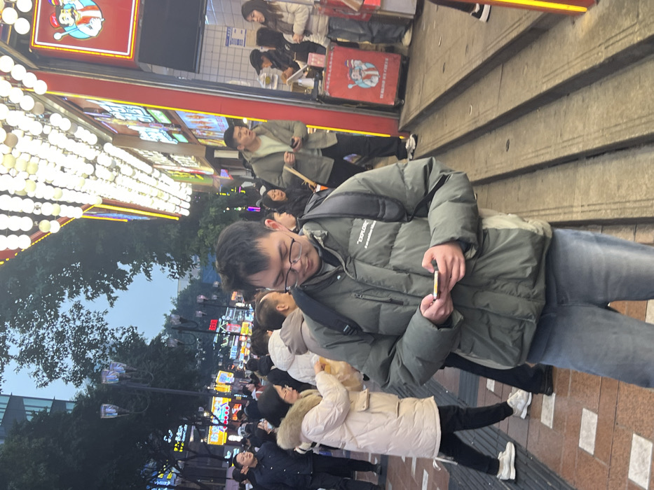 The image captures a bustling urban street scene in Yuzhong District, China, at dusk or early evening, illuminated by extensive artificial lighting.
In the foreground, a male individual of East Asian descent stands slightly off-center, looking down at a smartphone held in both hands. He wears a dark green puffer jacket with the text "EXPLORE" on the upper right chest, light blue jeans, and clear-framed eyeglasses. A dark backpack is worn over his jacket.
To the left of the primary subject, a dense crowd of pedestrians, predominantly East Asian, occupies a paved sidewalk. Many are dressed in winter clothing, including puffer jackets. A woman in a light-colored puffer jacket with a fur-lined hood is visible, holding a light-colored item, possibly food. In the background on the left, multi-globe streetlights stand among trees with visible branches. Buildings with various illuminated signs are present further down the street.
To the right of the primary subject, a brightly lit commercial building facade extends along the street. The upper section features numerous rows of spherical, white overhead lights. Below these lights, storefronts display vibrant neon signage with Chinese characters and cartoon figures, including one depicting a chef. A set of three concrete steps leads down from the street level to a shop entrance. A red vendor cart with a cartoon character and text is situated near the steps. Several individuals are visible near a counter within the shop entrance, some appearing to be customers or staff. One man in a light green jacket holds a smartphone and a slender stick-like object.