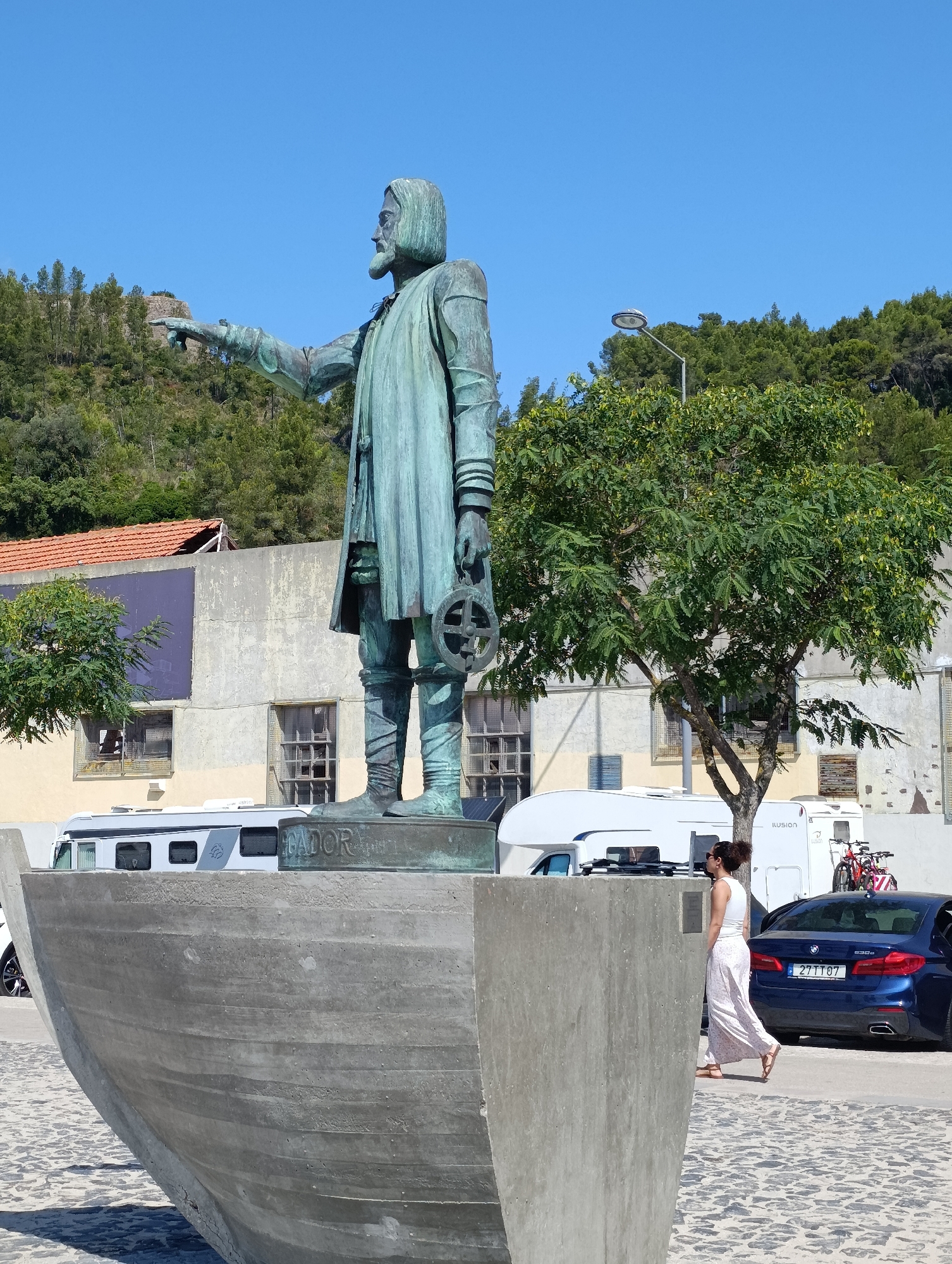 The image captures a bronze statue of a historical figure, possibly a navigator, standing on a concrete pedestal shaped like a boat. The statue depicts a man with a beard, wearing a long coat and boots, pointing forward with his left arm and holding a navigational instrument in his right hand. The base of the statue has the inscription "CCADOR".
In the background, a modern city scene unfolds. A woman in a white top and a long, floral-patterned skirt is walking towards the right of the frame. To her left, a dark blue BMW sedan is parked with its driver's side door open. Behind the statue and the woman, a white camper van and a red bicycle are visible. The environment appears to be a public space with a cobblestone ground. The backdrop features buildings with aged facades, some with reddish-tiled roofs, and a dense green hillside covered in trees under a clear blue sky.
This scene is located at the Monumento aos Descobridores in Setúbal, Portugal, which commemorates the Portuguese Age of Discovery. The monument is situated near the waterfront of the Sado River estuary.