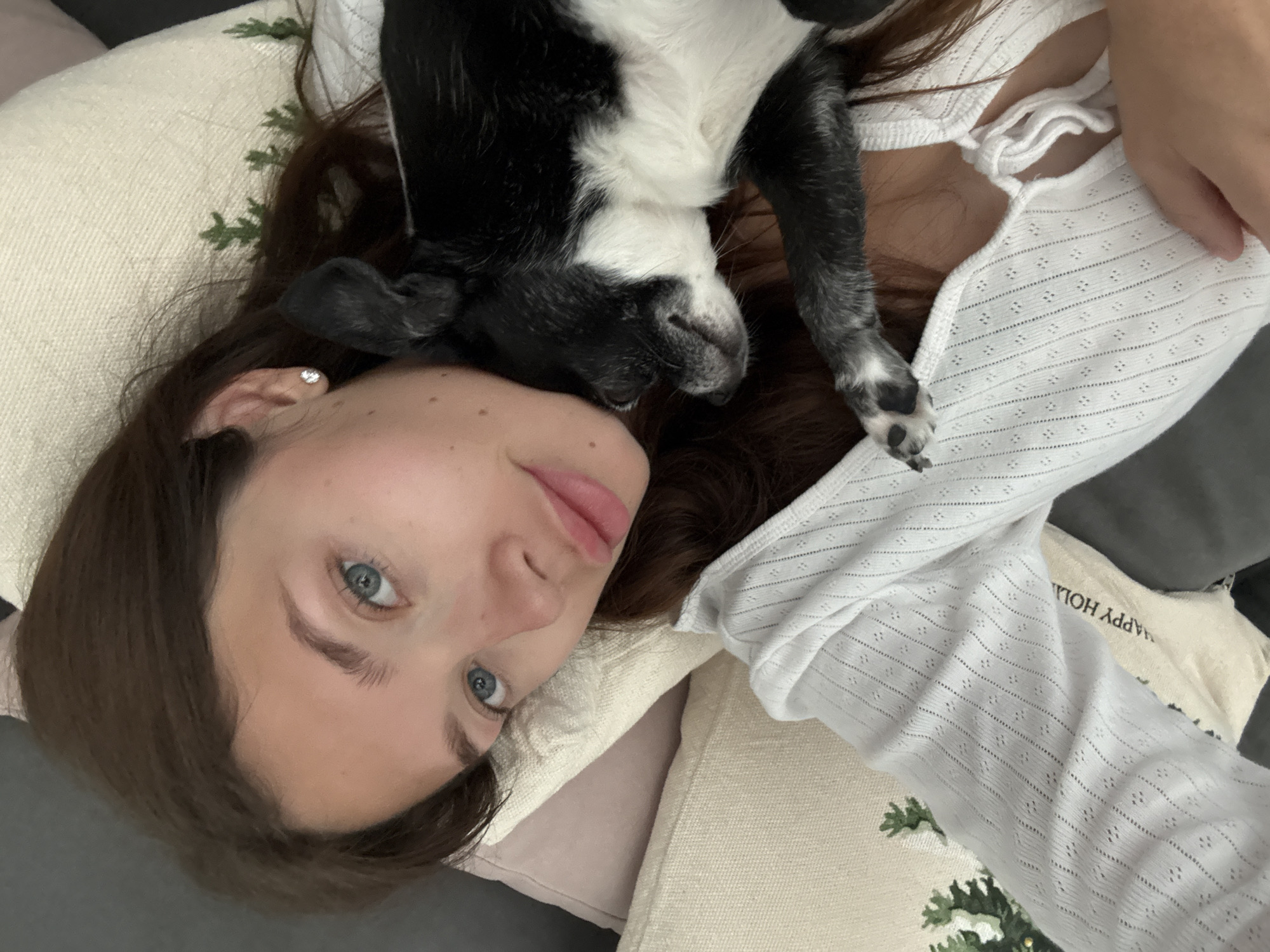 A young woman with fair skin and light blue eyes is lying on a sofa, looking up at the camera. Her brown hair is spread out on a pillow. A black and white dog is lying on her, with its head resting near her face and one paw near her chest. She is wearing a white, lacy top. The pillow beneath her head is light tan with green embroidery. There is a second pillow underneath with "HAPPY HOL" printed on it. The location appears to be an indoor setting in an unknown city.