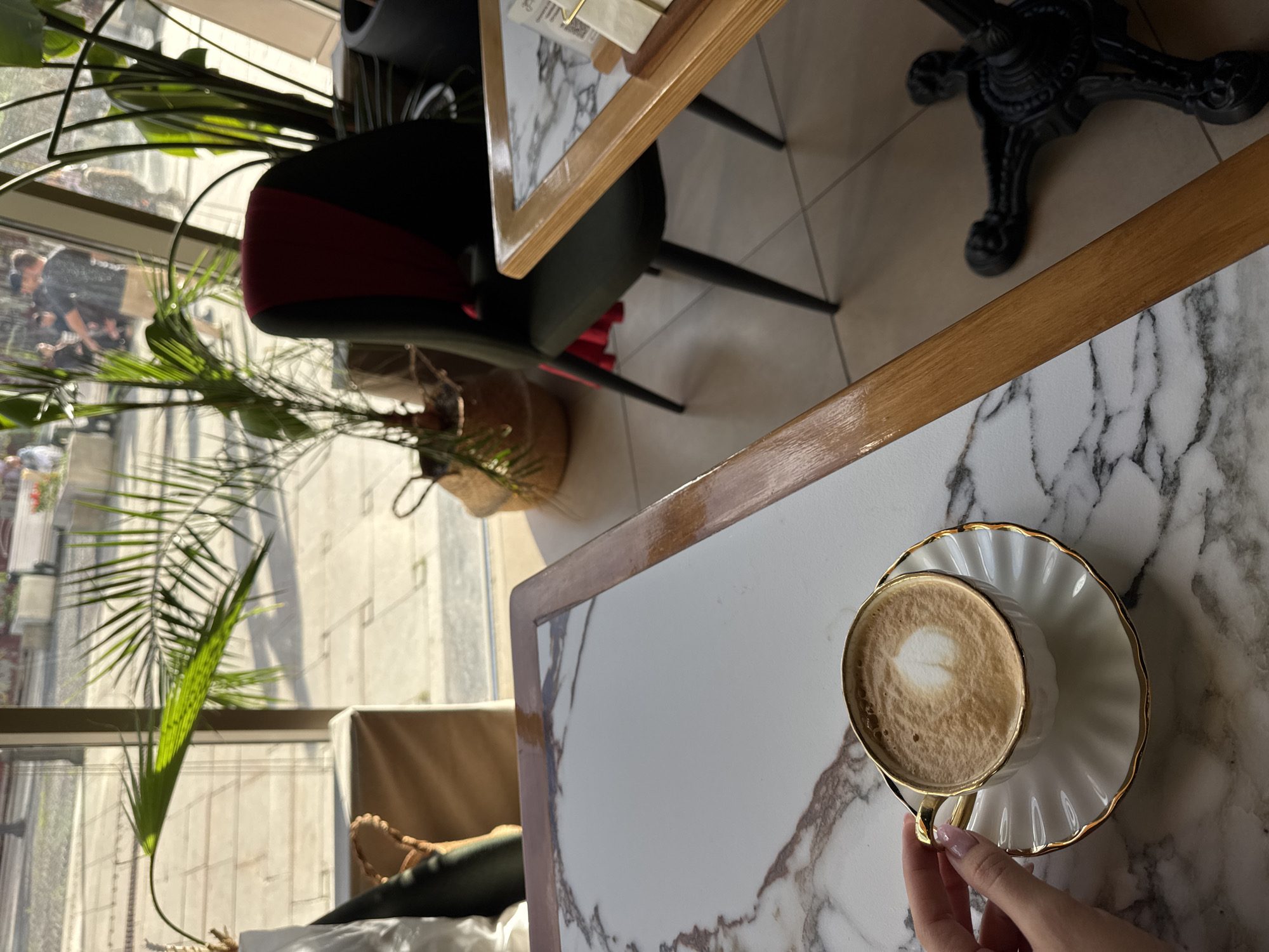 A close-up, eye-level shot shows a person's hand holding a gold-rimmed, white ceramic cup filled with cappuccino art. The cup sits on a matching saucer, both with a fluted edge. The coffee has a heart shape in latte art. The table surface is white marble with grey veins. In the background, through a large window, a street scene is visible with people walking and potted plants. One large potted palm is in the foreground on the left, casting shadows. A dark chair with a red accent is visible on the right, behind a second marble-topped table with a wooden frame. The floor is tiled in a neutral color.