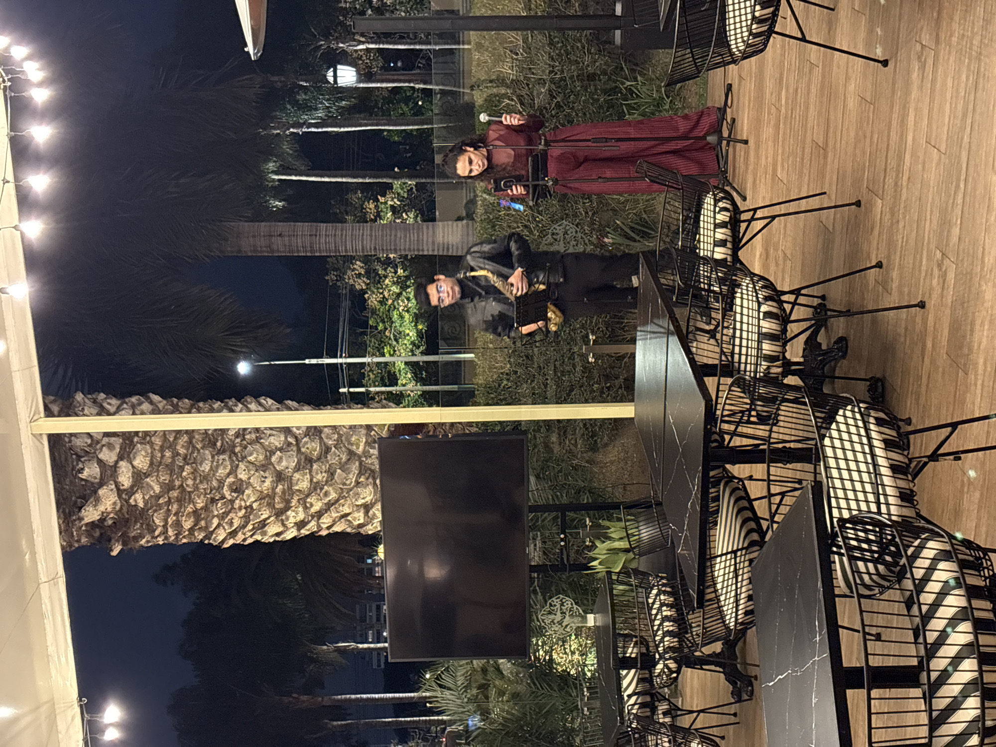 In San Isidro, Peru, at nighttime, a live performance is taking place in an outdoor dining area. Two performers are on a stage area. A man on the left is playing a saxophone, wearing a black leather jacket and black pants. A woman on the right is singing into a microphone, dressed in a dark red, wide-legged jumpsuit. They are positioned in front of a lush green hedge.
In the foreground, several tables with black marble tabletops and black metal chairs with striped black and white cushions are arranged on a wooden floor. The tables are set with place settings, suggesting a restaurant or event space.
To the left of the performers, a large, black flat-screen television is mounted on a stand. Behind the performers, palm trees and other foliage are visible. String lights are strung overhead, illuminating the scene. The overall atmosphere appears to be a sophisticated outdoor venue for dining and entertainment.