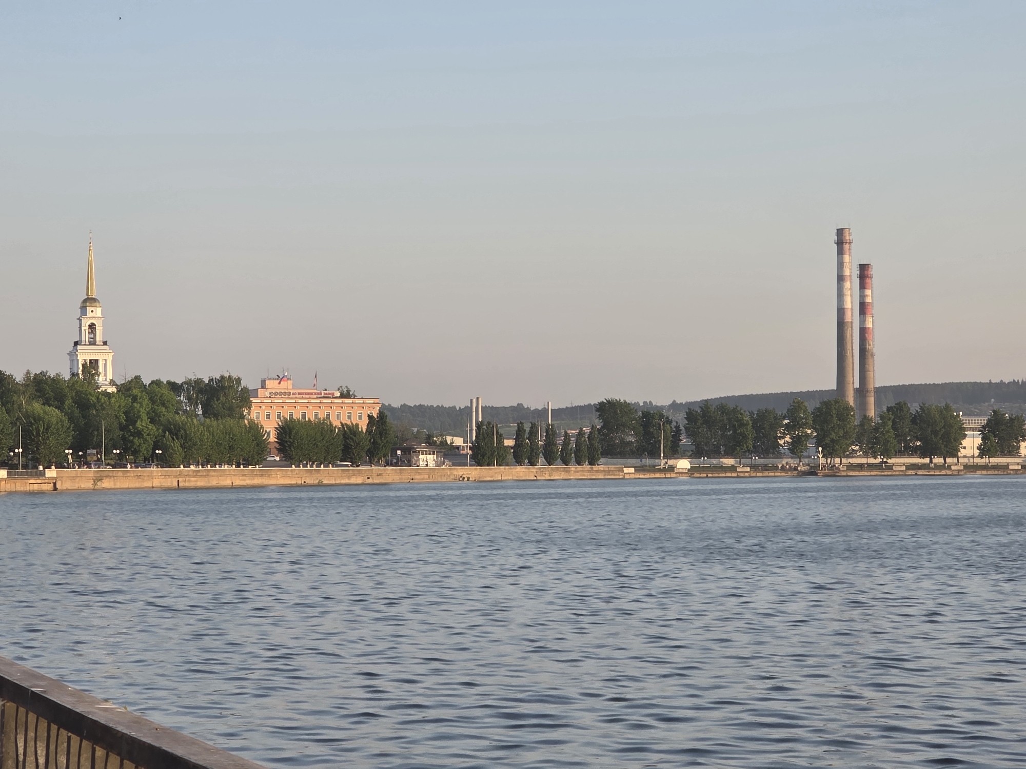 The image depicts a waterfront scene in Votkinsk, Russia. In the background, the cityscape includes a prominent church steeple, a multi-story brick building with Russian flags on top, and two tall industrial chimneys. The chimneys have alternating red and white stripes near their tops. A line of green trees separates the buildings from the waterfront. The foreground consists of a large body of water with gentle ripples on its surface. A partial view of a wooden railing is visible at the bottom left corner of the image.