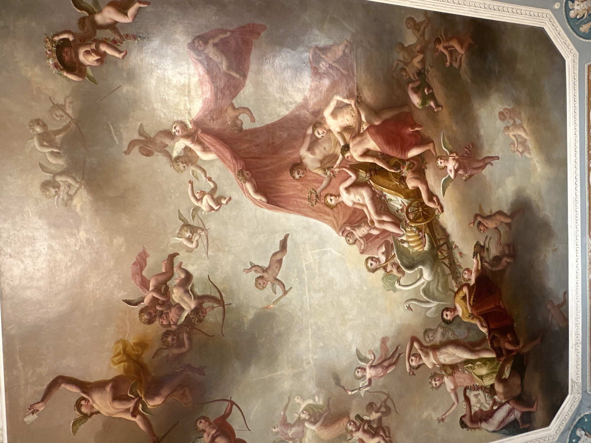 This is a ceiling fresco in the Grand Palace in Gatchina, Russia, depicting a mythological scene with numerous cherubs and figures in a cloudy, ethereal environment. In the upper left, a winged male figure with a helmet and a golden drapery is depicted mid-flight, holding an object in his outstretched hand. Numerous cherubic figures, some with wings, are scattered throughout the fresco, many holding bows and arrows, suggesting a theme of love or Cupid. One group of cherubs appears to be carrying a basket overflowing with flowers. In the center of the fresco, a large, flowing pink drapery is being held aloft, revealing several nude or semi-nude figures. Below this, a chariot is being pulled by two white swans, carrying several female figures adorned with flowers and laurel wreaths. To the right of the chariot, more cherubic figures are depicted, some appearing to be climbing or reaching. The overall impression is one of heavenly revelry and mythological grandeur, rendered in soft, warm tones against a backdrop of muted clouds.