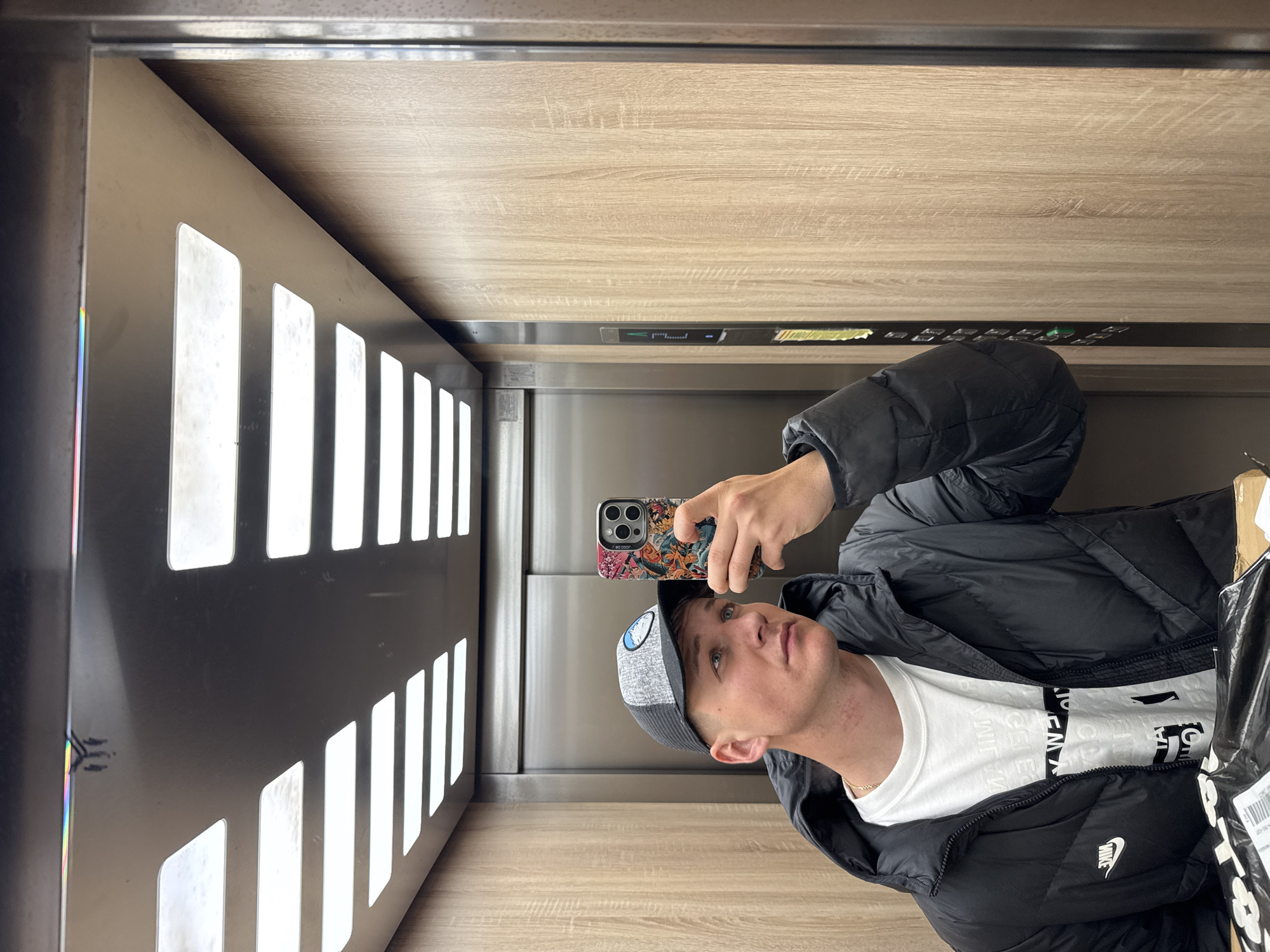 A man takes a selfie in an elevator in Plovdiv, Bulgaria. He is wearing a black puffer jacket, a white t-shirt, and a grey and black baseball cap. He is holding a phone with a colorful case. The elevator has metal walls with vertical rectangular cutouts on one side and light wood paneling on the others. There is a panel of buttons above his head.