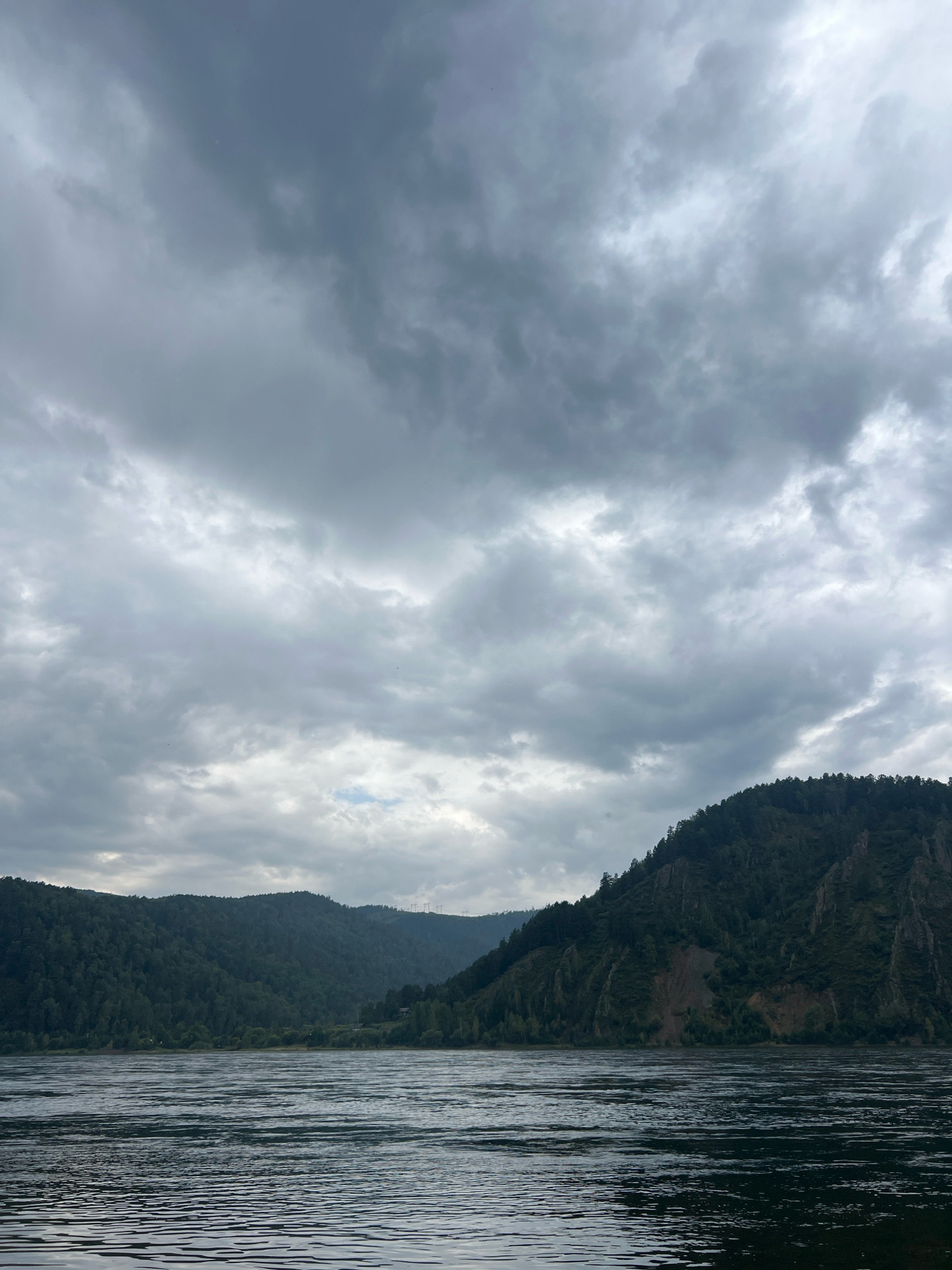 The image depicts a wide river in the foreground, with dark, choppy water reflecting the overcast sky. Beyond the river, a dense, dark green forest covers rolling hills. The hills are steep and rocky in some areas, with sparse vegetation clinging to the slopes. The sky is filled with heavy, gray clouds, suggesting a gloomy or possibly stormy day. Power lines are visible in the distance on the right side of the frame, indicating human presence and infrastructure. The overall scene is natural and somewhat imposing, dominated by the vastness of the water and the dark, forested landscape under a dramatic sky. This landscape is characteristic of the area around Divnogorsk, Russia, known for its natural beauty along the Yenisei River.