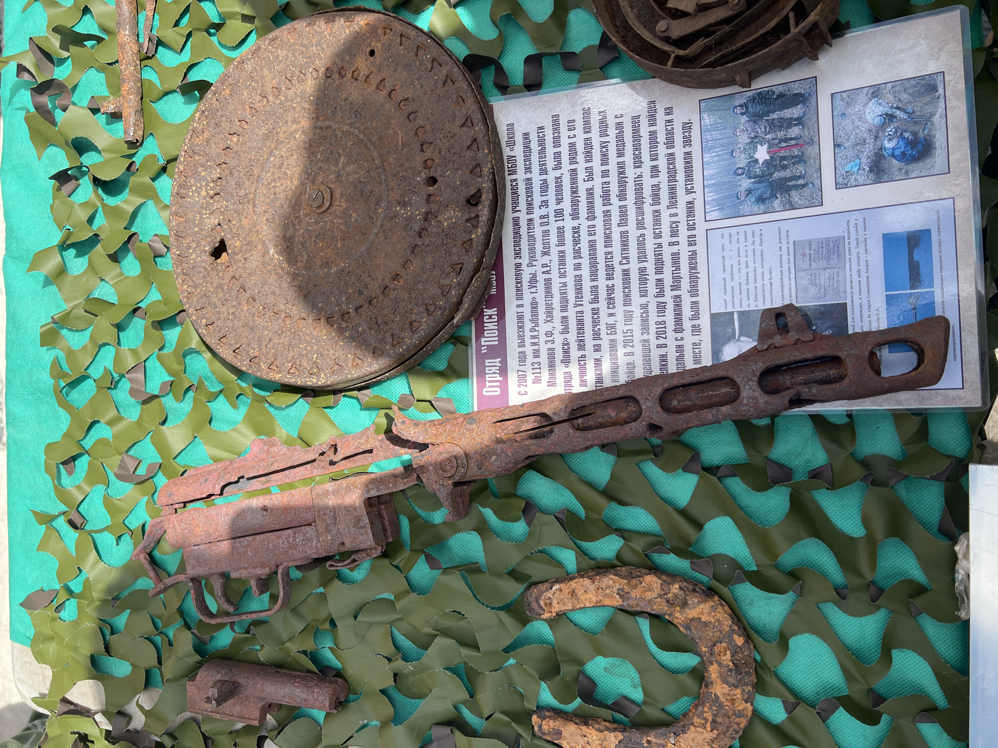 The image shows an exhibit, likely in Ufa, Russia, related to a search expedition group. Dominating the scene is a heavily rusted gun, possibly a submachine gun, resting on a green camouflage net. Other artifacts include a corroded circular metal plate (possibly a manhole cover) and a rusty horseshoe-shaped object. Behind these items is a sheet with text in Russian describing the "Poisk" (Search) squad, operating since 2007 by students of School #113 named after I.I. Rybalko in Ufa. The text details the squad's activities, including finding the remains of over 100 people, identifying a lieutenant by a comb with his name scratched into it, and discovering a medallion with an inscription in 2015. It also mentions finding the remains of a soldier with a medallion named Martynov in 2018 in the Leningrad region. There are also photos on the sheet depicting the search squad, the process of finding remains, and the memorial star placed in the forest where the remains were found.