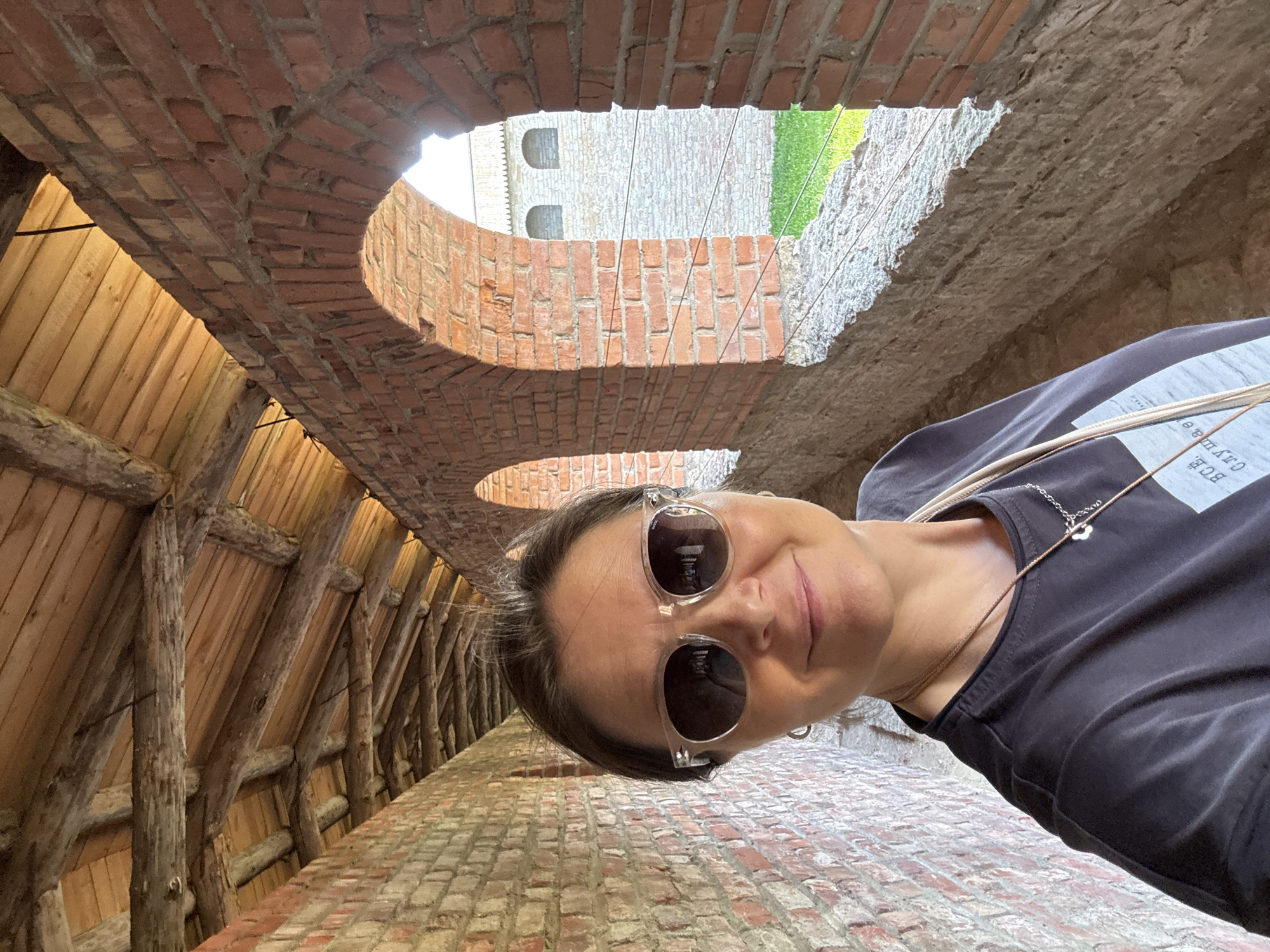 The image shows a woman taking a selfie inside the Oreshek Fortress in Shlisselburg, Russia. She is wearing sunglasses and a dark t-shirt. The fortress walls are made of brick, and the ceiling is constructed with wooden beams and planks. Through arched openings in the wall, a grassy area and another building with arched windows can be seen.
