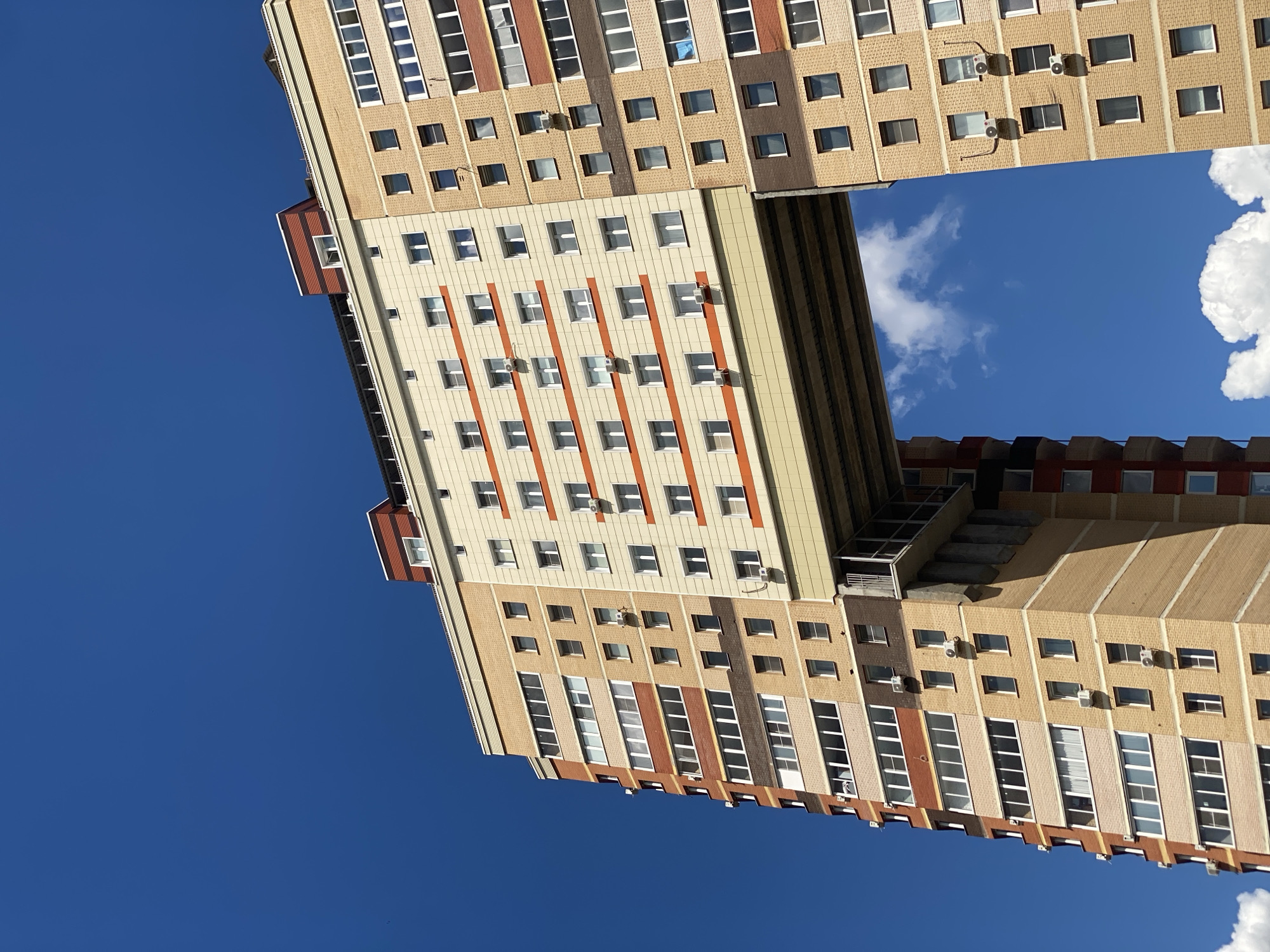 This media file is a low-angle, close-up shot of a modern, multi-story apartment building against a clear blue sky with scattered white clouds. The building's architecture is characterized by a repeating pattern of beige and light-colored facade panels, accented by vertical orange stripes on some sections. Numerous windows are visible across the building's facade, many of which are white-framed and reflect the bright sky. Several external air conditioning units are mounted on the walls.
The image appears to have been taken on a sunny day, likely in the daytime, given the clear sky and bright lighting. The composition is unusual as the building is shot from a very low perspective, making it seem to stretch upwards into the sky. The building's structure is somewhat segmented, with different sections appearing to be connected or juxtaposed. One section features a prominent red-roofed extension. The overall impression is one of urban architecture bathed in natural light. No people are visible in the frame, and there is no visible text. The location context is identified as Kotelniki, Russia.