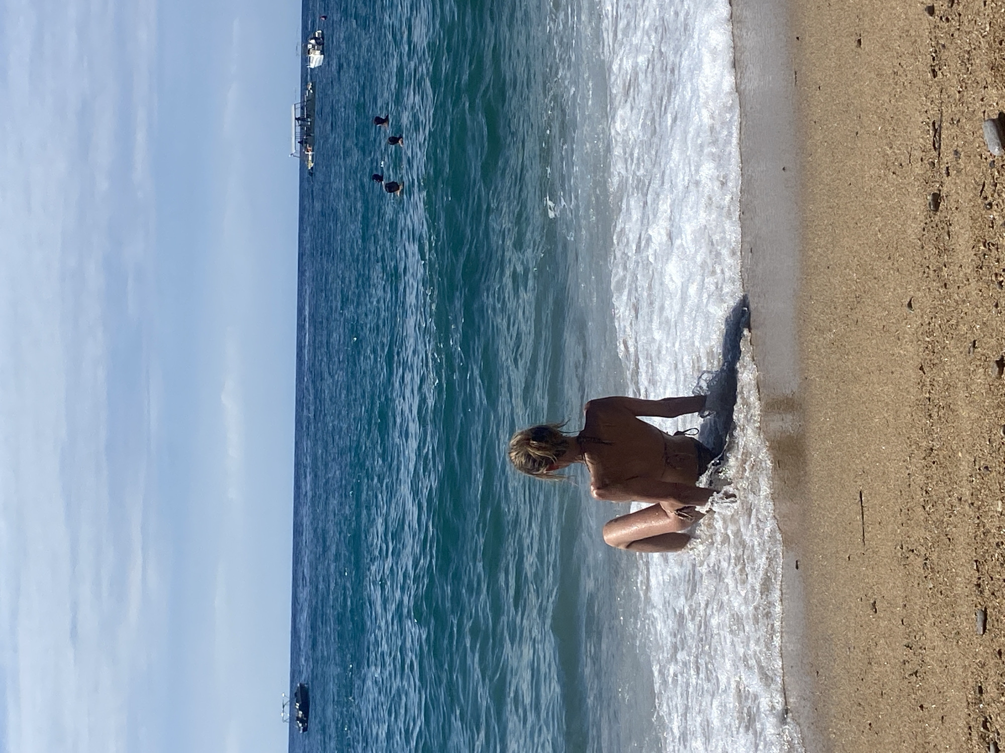 The image captures a vibrant daytime scene at the beach, looking out over the water from the shoreline. In the foreground, a person with blonde or light brown hair, appearing to be female and wearing a swimsuit, crouches at the water's edge, interacting with the foamy white surf as waves wash ashore. The individual's back is partially visible, facing towards the sea.
The sandy beach, interspersed with small pebbles, stretches to the right. The calm, blue-green sea extends across the middle ground, showing gentle ripples on its surface. Further out in the water, several dark, round buoys are visible, along with multiple boats and ships, including at least one larger vessel and a smaller motorboat. The horizon line is visible in the distance, separating the sea from the clear, bright blue sky above, which shows only faint cloud formations.
The scene suggests a sunny day with pleasant weather, ideal for a beach outing. The setting is consistent with a coastal environment, specifically in Sveti Stefan, Montenegro. There is no visible text in the image.
