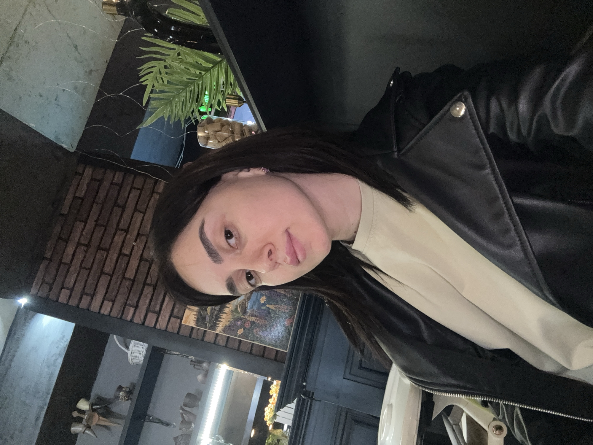 A person is photographed indoors, likely at a restaurant or cafe in Tbilisi, Georgia. The person has dark hair and is wearing a light-colored shirt under a black leather jacket. Behind the person is a brick wall and a display case with decorative objects. Plants and other decorative elements are also visible.