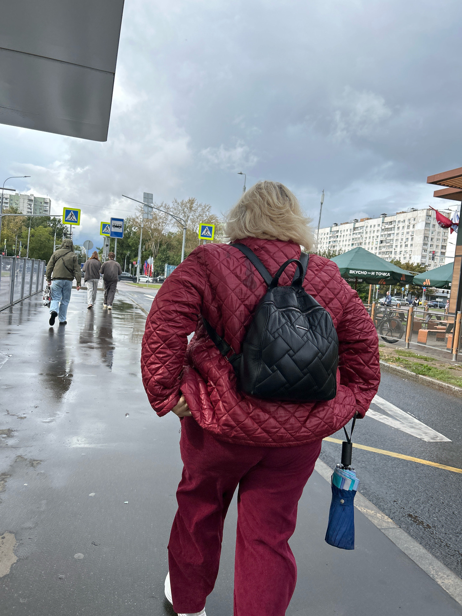 A blonde woman, seen from behind, walks on a wet sidewalk in Moscow, Russia. She wears a quilted red jacket and maroon pants, carrying a black backpack and a folded blue umbrella attached to her belt loop. Three men walk ahead of her on the sidewalk. In the background, there are apartment buildings, trees, street signs, and a fast-food restaurant with green umbrellas. The sky is overcast with clouds.