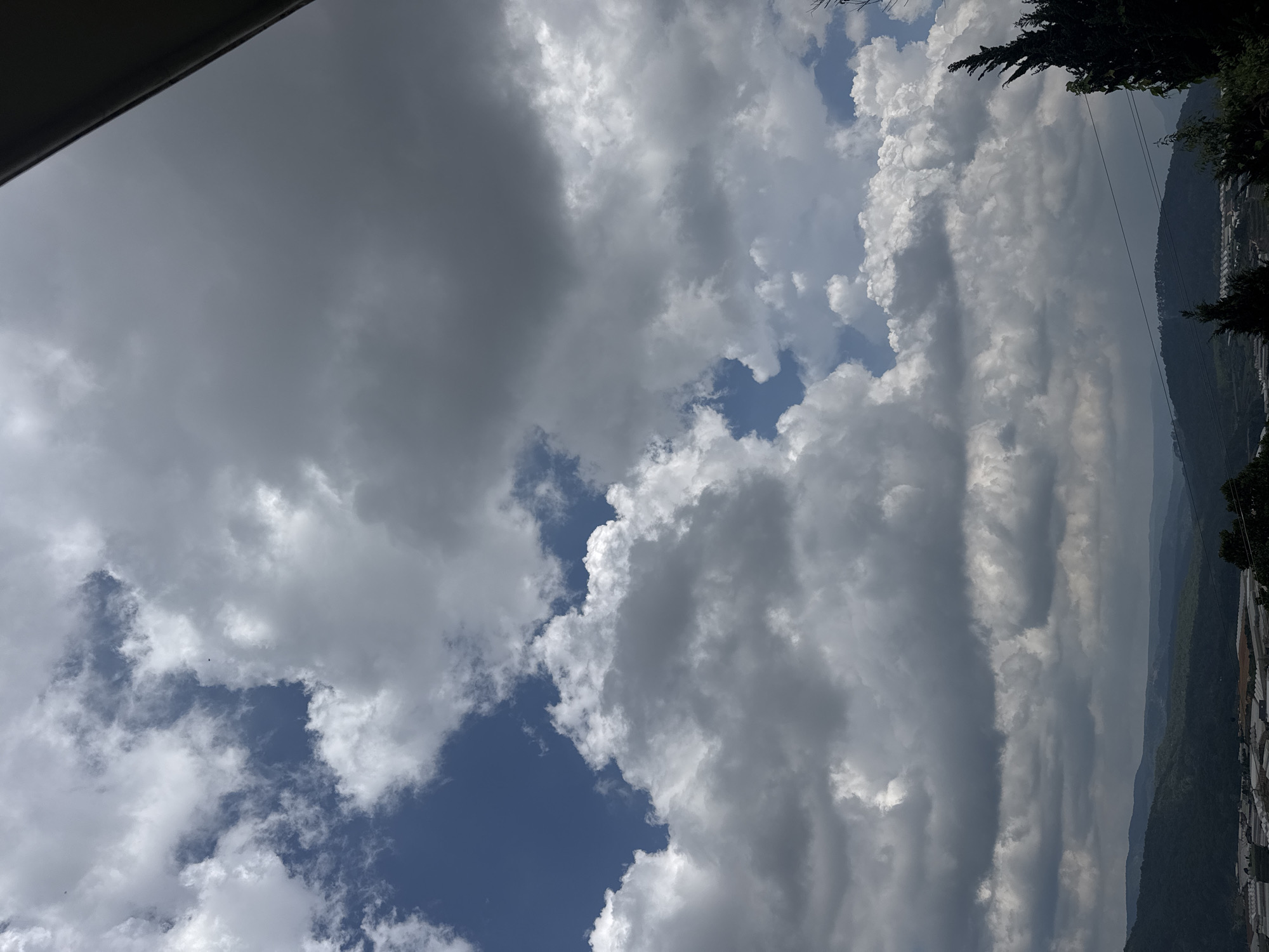 The image is a vertical shot of a partly cloudy sky over a landscape in Đa Lộc, Vietnam. The sky is filled with a mix of fluffy white cumulus clouds and darker, more diffused grey clouds. Patches of bright blue sky are visible between the clouds. In the foreground, the top edge of a dark structure, possibly a roofline or overhang, is visible in the upper right corner. The middle ground shows rolling hills covered in dark green vegetation. In the lower portion of the image, a cluster of greenhouses with white roofs and frames can be seen, indicating agricultural activity. A few dark green evergreen trees are visible along the right edge and scattered throughout the landscape. The overall impression is of a bright, overcast day with a hint of agricultural development in a hilly region.