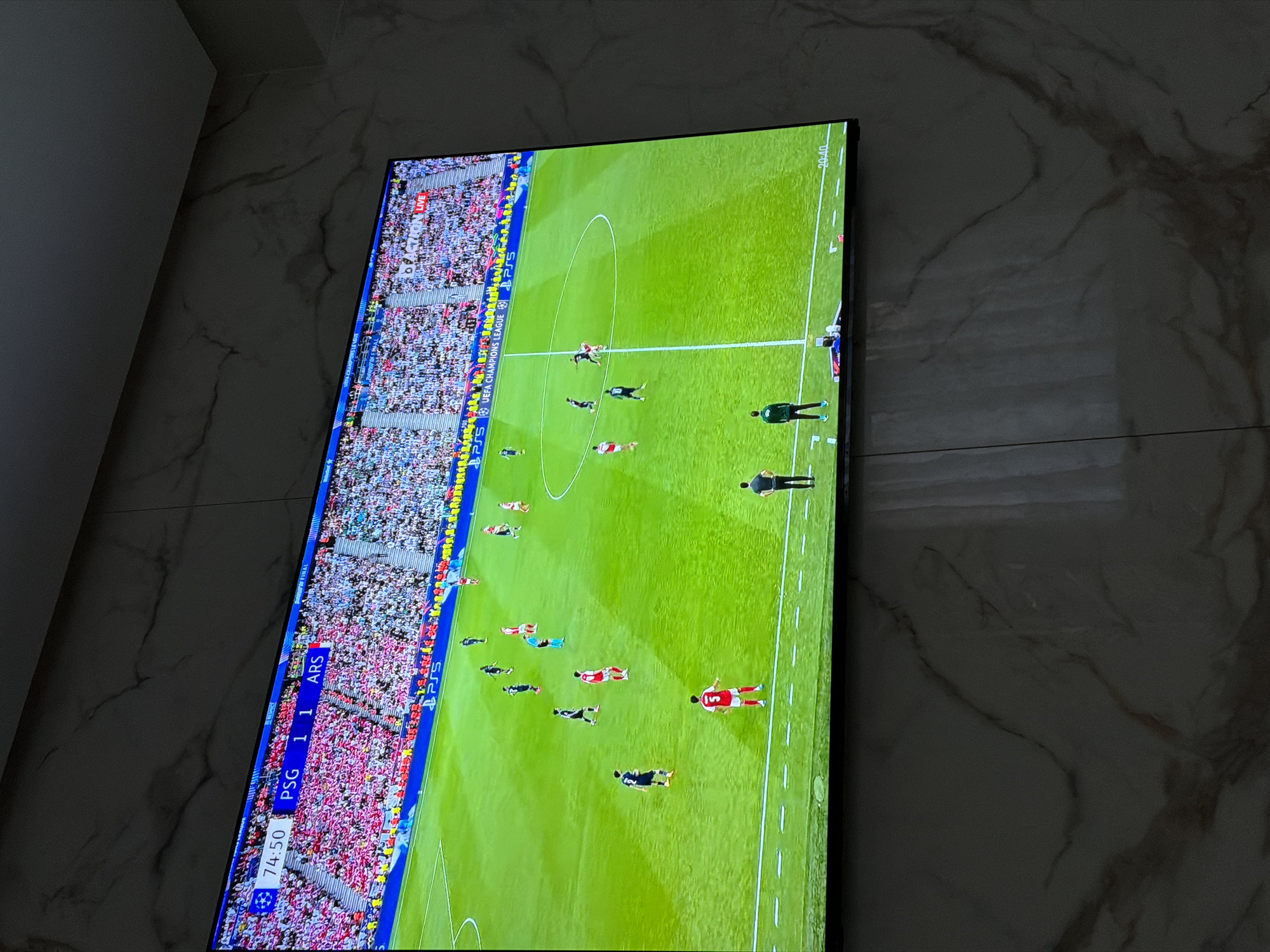The image shows a television screen displaying a live soccer match between Paris Saint-Germain (PSG) and Arsenal (ARS). The score is 1-1 at 74 minutes and 50 seconds into the game. The game appears to be a UEFA Champions League match. The view of the field shows players, stadium seating, and referees. The location is not present in the image.