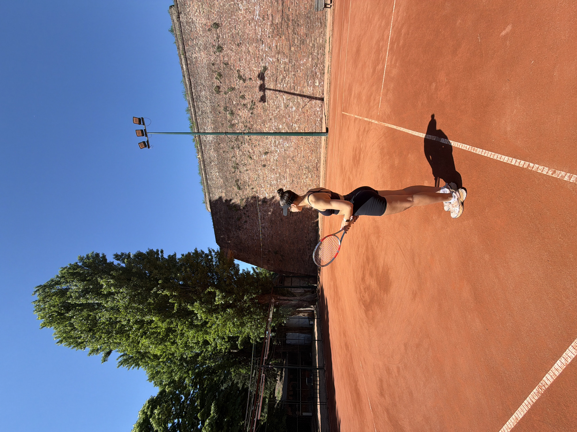 The image captures a moment on a tennis court in Belgrade, Serbia. A woman dressed in black sportswear and white sneakers stands on the reddish-brown clay court, holding a tennis racket. The court lines are clearly visible. Behind her, a high brick wall, partially covered with greenery, forms a backdrop, suggesting the presence of a historical structure or fortification. A tall green lamppost with three lamps extends upwards beside the wall, casting a shadow onto the court. A mature, leafy green tree is visible on the left side of the frame. The sky is a clear, bright blue. The scene is bathed in sunlight, casting distinct shadows.