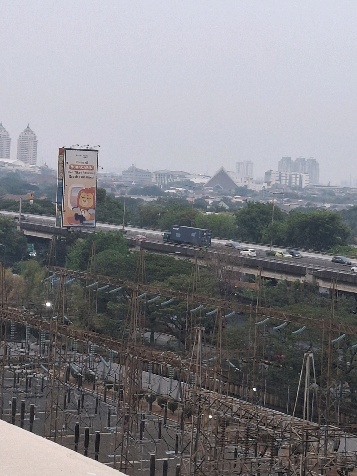 The image shows a hazy, overcast sky over a city skyline. In the foreground, there is an electrical substation with numerous insulators and conductors. A highway overpass runs through the middle ground, with several vehicles visible, including cars and a truck. A large billboard is prominently displayed on the left side of the overpass, featuring an advertisement for "BOOKCABIN" with an image of a cartoon lion. In the background, a dense urban landscape with various buildings, including high-rises and a pyramid-shaped structure, is visible. The overall impression is of a bustling urban environment under a muted sky. The specific location is Central Jakarta, Indonesia, as indicated by the presence of the electrical substation, typical infrastructure, and architectural styles common in the region. There are no identifiable people in the image. The scene depicts a static urban environment with ongoing traffic on the highway.