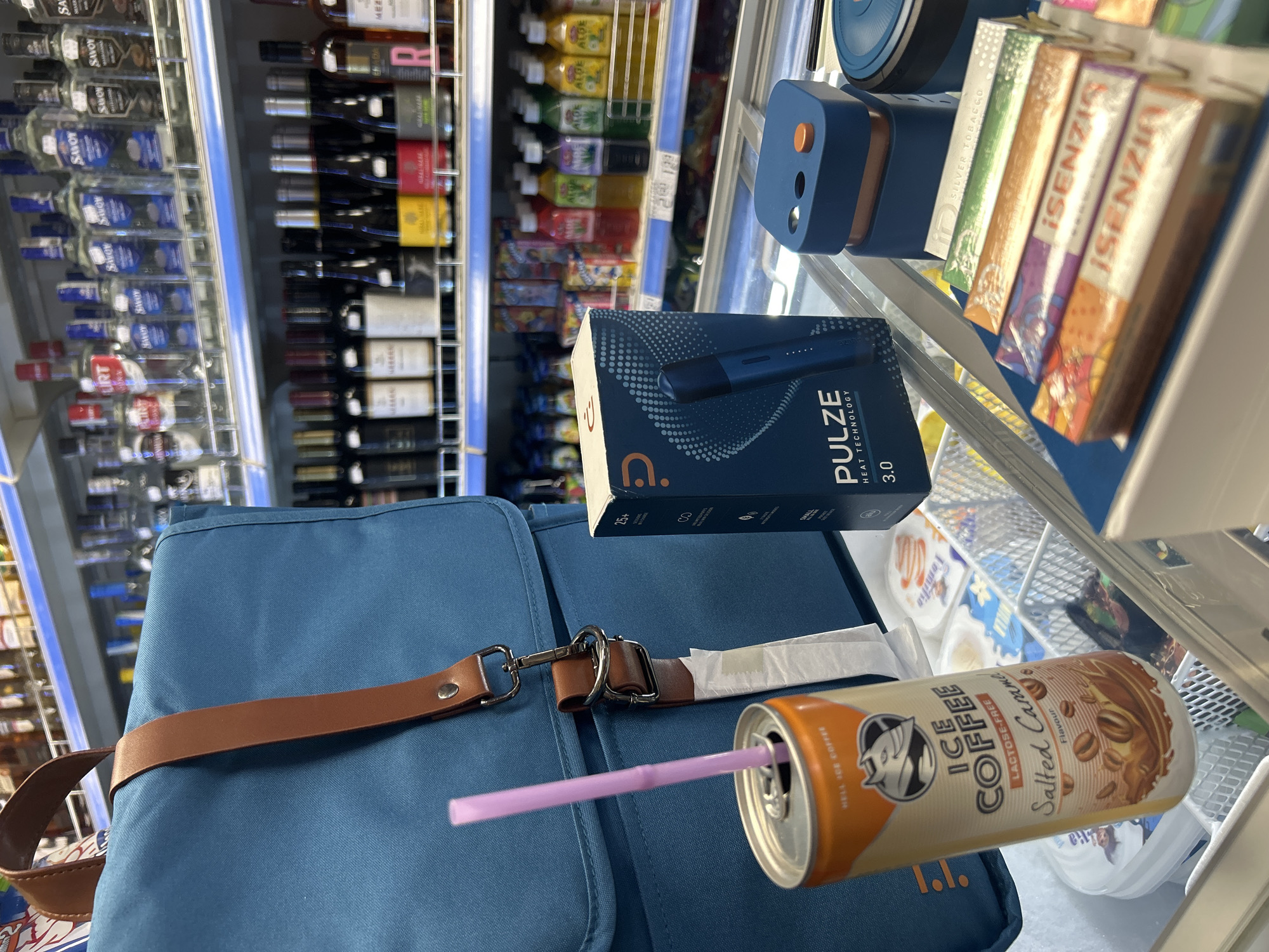 The image shows a close-up of several items on display in what appears to be a convenience store or a shop with refrigerated sections. In the foreground, a teal-colored bag with brown leather straps is partially visible. To the right of the bag is an orange can of "ICE COFFEE Lactose-Free Salted Caramel Flavour" with a pink straw inserted into it. Next to the can, a blue and black box with "PULZE HEAT TECHNOLOGY 3.0" printed on it is displayed. To the right of the box is a small blue charging dock with an orange button and a light. In the background, shelves are stocked with various bottles of alcohol, including what looks like vodka and wine, as well as other products in colorful packaging. The overall lighting suggests an indoor setting, possibly a retail environment. There are no people visible in the image.