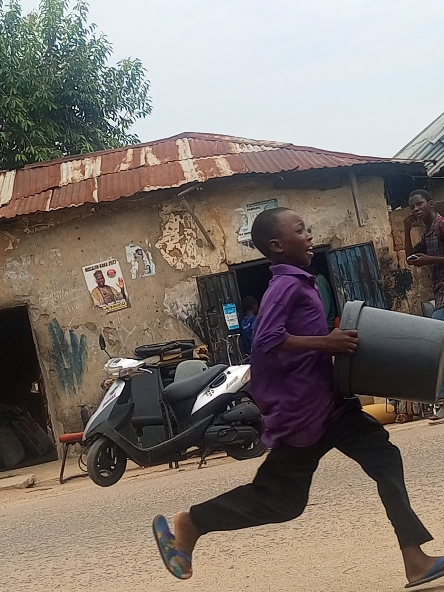 A young boy in a purple shirt and black pants is running on a paved road carrying a large, gray bucket. He is wearing blue sandals. Behind him is a building with a rusty, corrugated metal roof. The building's walls are peeling and have posters on them, including one that says "Magajin Kaka 2027". A white and black scooter is parked in front of the building. A man is standing in the doorway of the building, looking at a mobile phone. There are other people in the background, near the building entrance. The sky is overcast. The image appears to be taken in a rural or low-income urban area, likely in Maiburiji, Nigeria, based on the text on the poster.