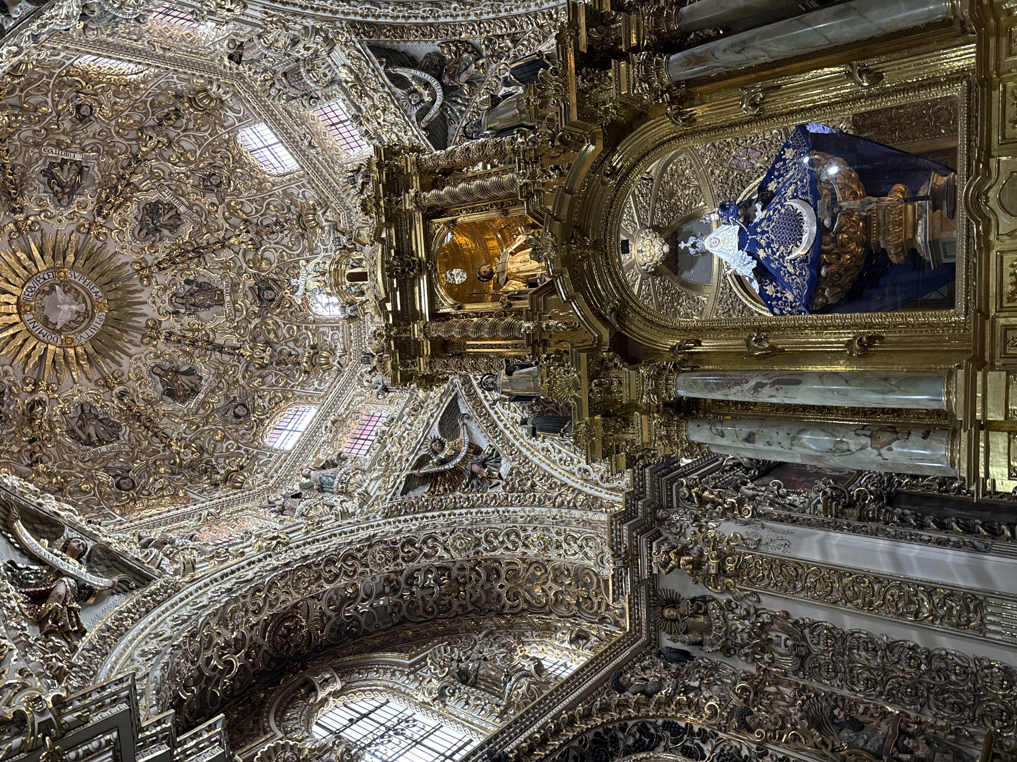 This media file captures the opulent interior of a grand religious building in Puebla, Mexico, showcasing a magnificent vaulted ceiling and a multi-tiered altarpiece. The architectural style is characterized by the elaborate Baroque or Churrigueresque tradition, heavily featuring gold leaf and intricate stucco work.
The ceiling is a dense tapestry of white and gold, adorned with swirling patterns, floral motifs, and numerous small sculptures of angels, cherubs, and saints. Several windows are visible, allowing natural light to illuminate the space. A large, central golden sunburst design on the ceiling features inscriptions in Latin: "REQUIESCIT IN DOMINI" and "SPIRITUS SUPEREUM." Other figures embedded in the ceiling hold scrolls with Latin phrases representing the gifts of the Holy Spirit, such as "SPIRITUM FORTITUDINIS," "SPIRITUM SCIENTIAE," "SPIRITUM CONSILII," "SPIRITUM TIMORIS DOMINI," "SPIRITUM PIETATIS," "SPIRITUM INTELLECTUS," and "SPIRITUM SAPIENTIAE."
To the right, a massive, gilded altarpiece (retablo) rises, covered entirely in gold leaf and detailed carvings. Within its upper golden niche, a figure, possibly a saint, is illuminated by a warm, golden glow. Below, a prominent statue of the Virgin Mary is displayed within a glass-enclosed section. She is depicted wearing a richly embroidered blue robe, a crown, and stands upon a crescent moon. A crucifix is also visible alongside her.
The scene conveys an overwhelming sense of grandeur and intricate craftsmanship, typical of colonial religious art in Mexico. No specific activity or event is taking place, as the image focuses on the architectural and sculptural splendor. The natural light filtering through the windows suggests it is daytime.