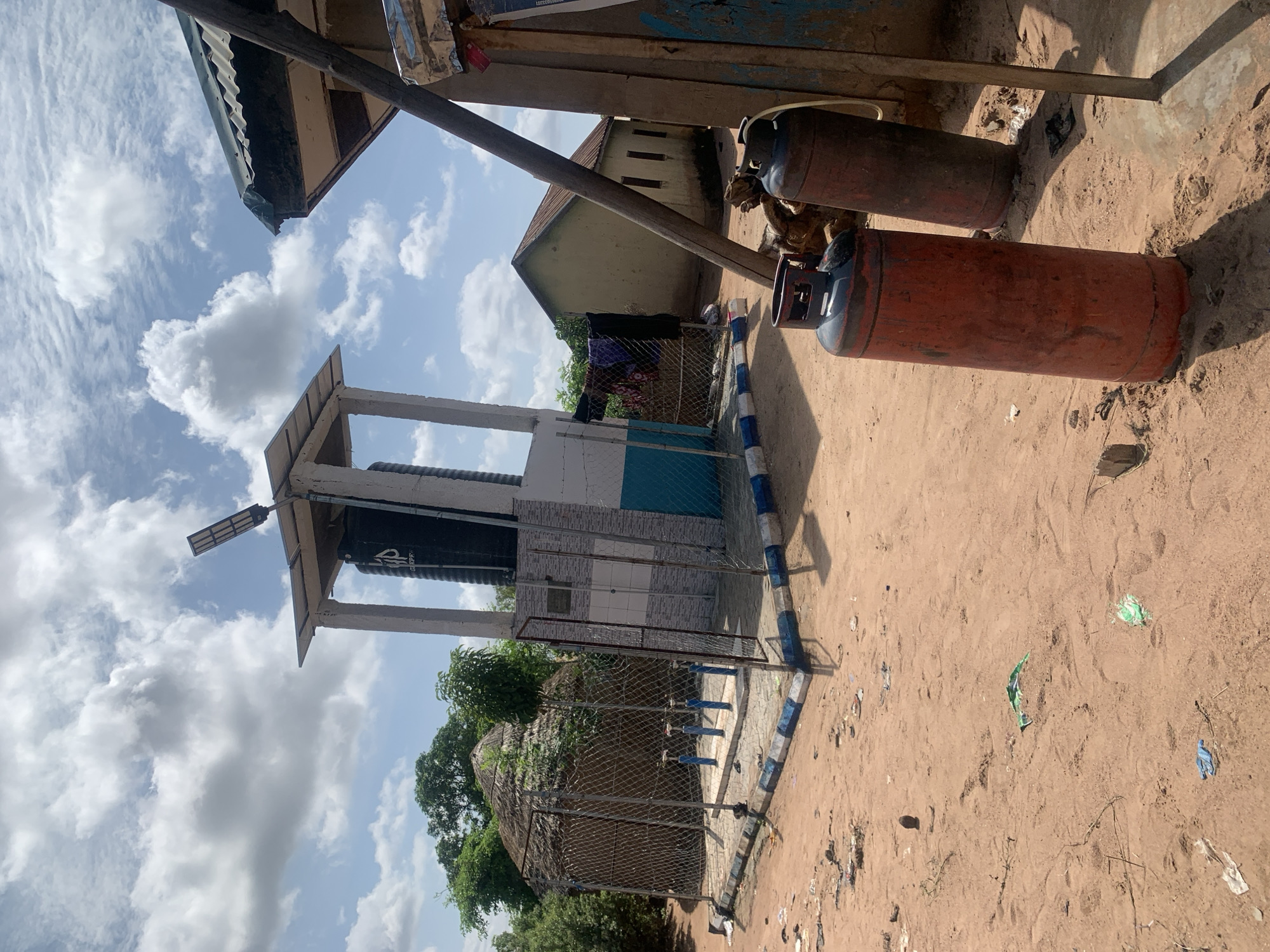 The image depicts a scene in Jalingo, Nigeria, featuring a water dispensing station and surrounding structures under a cloudy sky. In the foreground, two large red and brown propane gas cylinders stand upright on sandy ground, casting shadows. To the left, a water dispensing station is enclosed by a chain-link fence with blue and white striped edging along its base. Inside the fence are several blue water taps. A large black water tank is elevated on a concrete structure behind the taps, topped with a solar panel and light fixture. Adjacent to the water station is a small hut with a thatched roof. To the right, a part of a building with a corrugated roof is visible, with laundry hanging on a line. Further in the background, a pale yellow building with a reddish-brown roof stands amongst trees. The ground is sandy and littered with various debris, including plastic wrappers and a brown circular object that could be a stone or dried fruit.