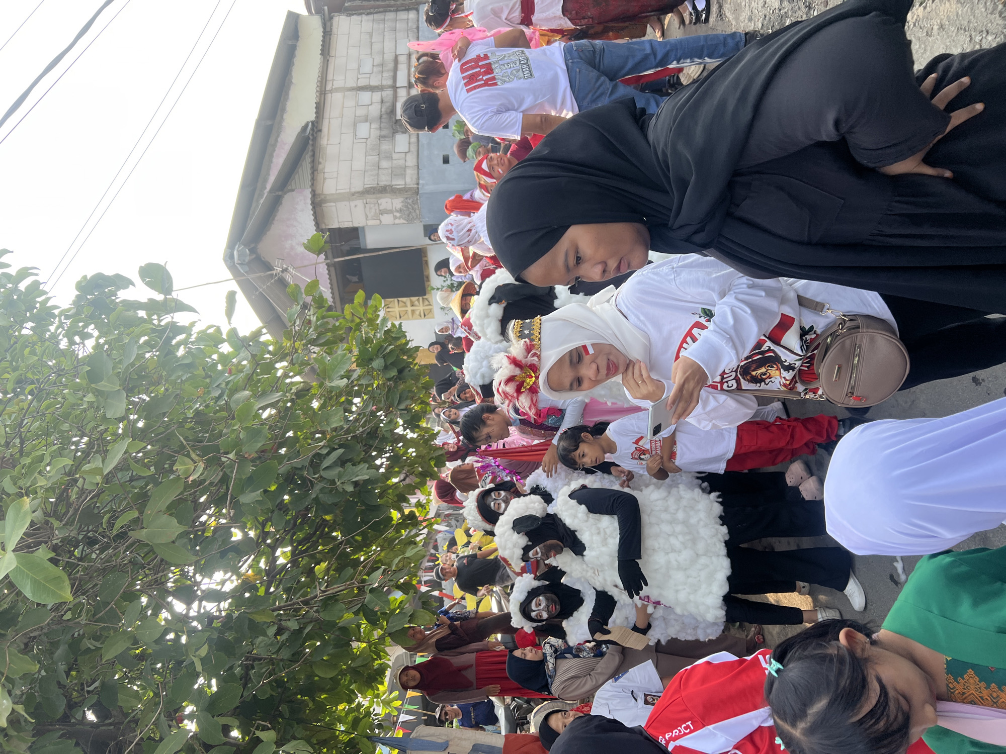 The image depicts an outdoor gathering of people, likely a parade or festival, in Klapanunggal, Indonesia. The scene is populated by individuals of various ages, many of whom are dressed in festive attire. In the foreground, a woman in a black hijab is looking to her right, with her hand on her hip. To her left, a young girl with green clothing and a pink scarf is looking down at her phone, and a child in a white hijab is visible. Behind them, several people are dressed in costumes resembling sheep, complete with white fluffy bodies and black faces painted with white makeup. Some participants are wearing white shirts with red and white decorations, suggesting a patriotic theme, possibly related to Indonesian Independence Day. A man in a white t-shirt with Indonesian text and jeans is walking away from the camera. In the background, there are buildings, trees, and power lines, indicating an urban or semi-urban environment. The overall atmosphere appears lively and communal.