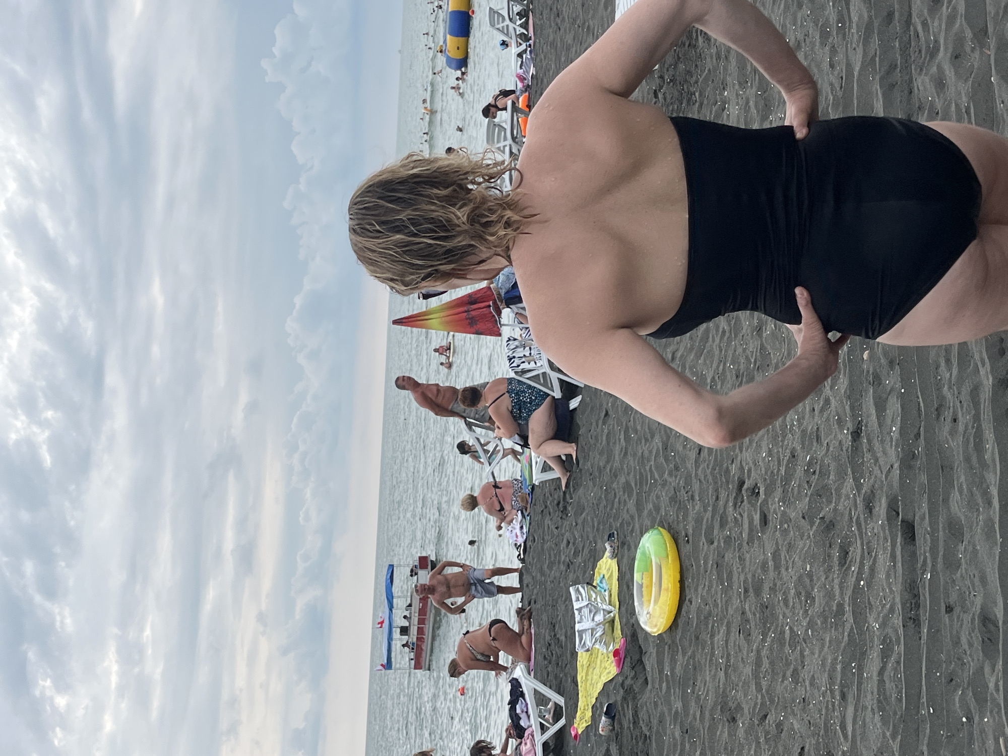 A medium shot captures a woman from behind, standing on a dark sand beach in Ureki, Georgia. She wears a black strapless swimsuit and has her hands on her hips, facing away from the camera towards a crowded beach. In the background, several people are seated on lounge chairs or walking near the water's edge. The sea is visible, with a red and blue boat and an inflatable water structure in the distance. The sky is overcast with clouds. In the foreground, a yellow and green inflatable ring lies on the sand.