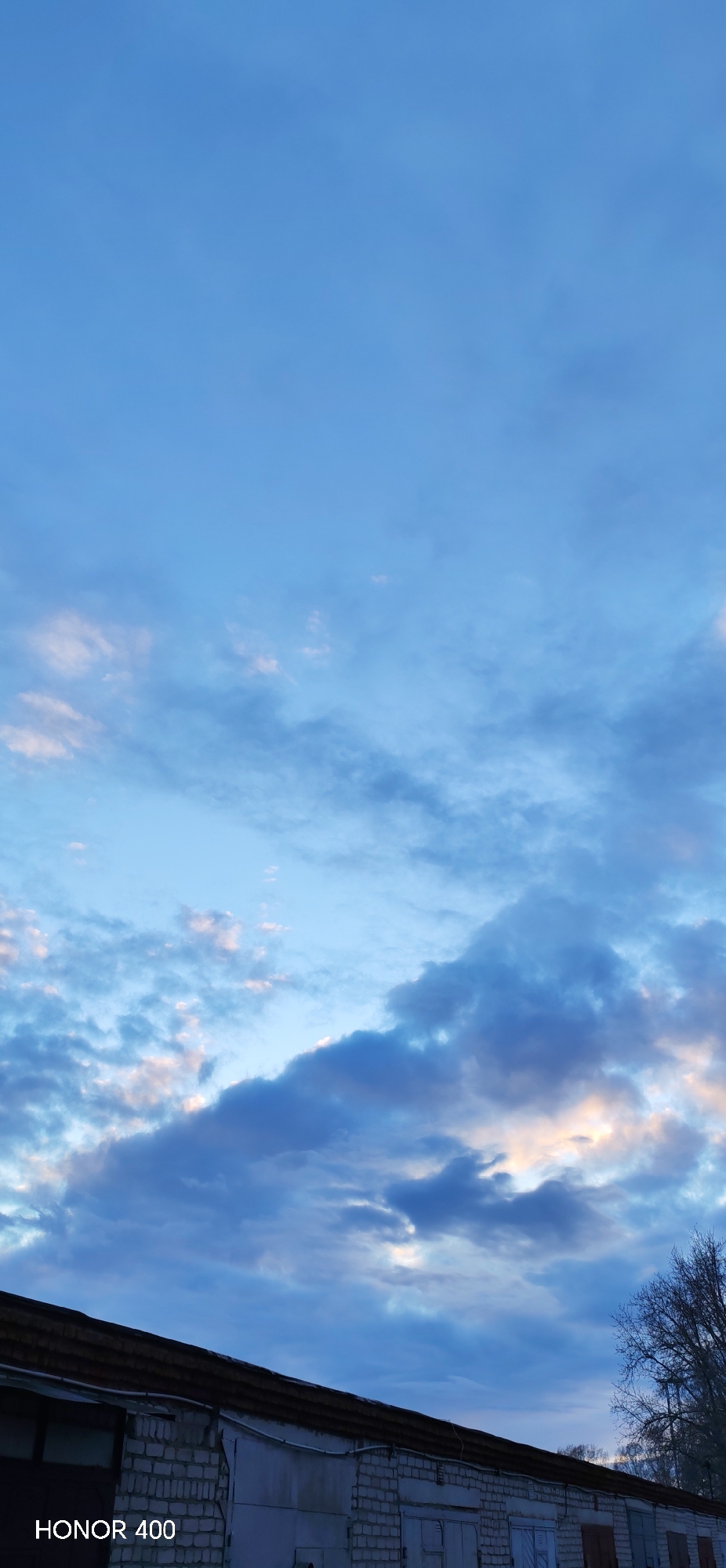 The image presents an outdoor scene in Chaikovsky, Russia, primarily featuring a vast sky above a row of buildings. The sky is a prominent element, displaying shades of blue, from a light, clear cerulean at the top to deeper, more overcast blues towards the horizon. Various clouds are scattered across the sky; some are light and wispy, touched with soft pink and golden hues, suggesting either sunrise or sunset. Other clouds are denser and darker, particularly towards the lower right portion of the sky.
In the bottom third of the frame, a series of low-rise buildings are visible, resembling garages or storage units. They are constructed with light-colored brick and feature multiple dark-colored doors or metal shutters. The roofs are dark and appear to be made of corrugated material. To the far right, the bare branches of a tree extend into the frame, indicative of a colder season such as late autumn or winter. The overall lighting is dim and ambient, creating a serene and somewhat melancholic atmosphere.
There are no people or active events taking place. The scene is static, capturing a quiet moment. In the bottom left corner of the image, the text "HONOR 400" is visible in white lettering.