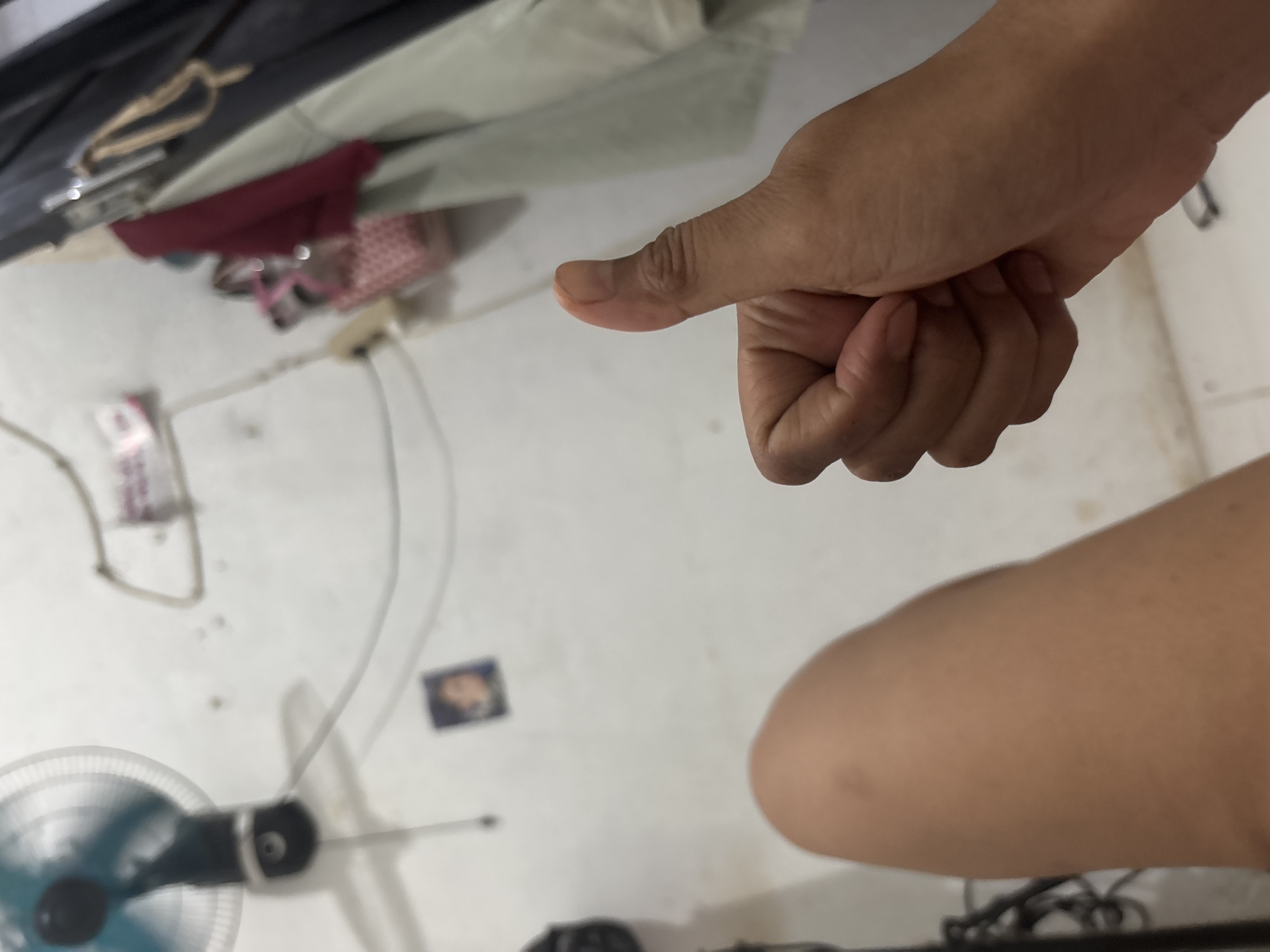 A hand gives a thumbs-up gesture in front of a person's leg. The background is a blurry interior scene with a wall fan on the left, some hanging clothes on the right, and a small framed picture on the wall. The image is taken from a low angle.